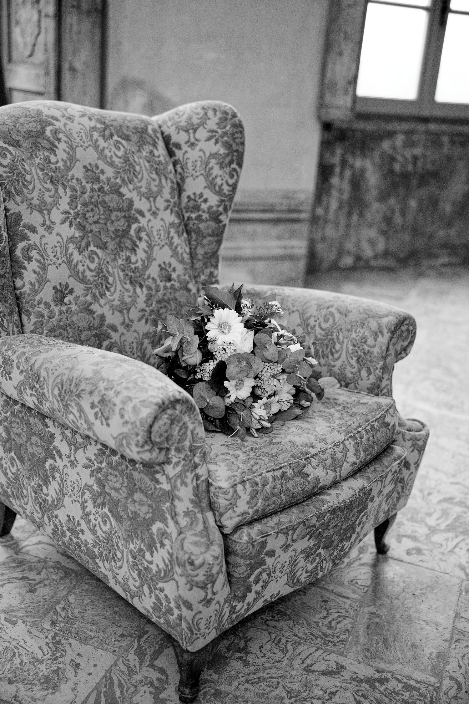 A black and white photo of a floral bouquet resting on a vintage floral armchair.