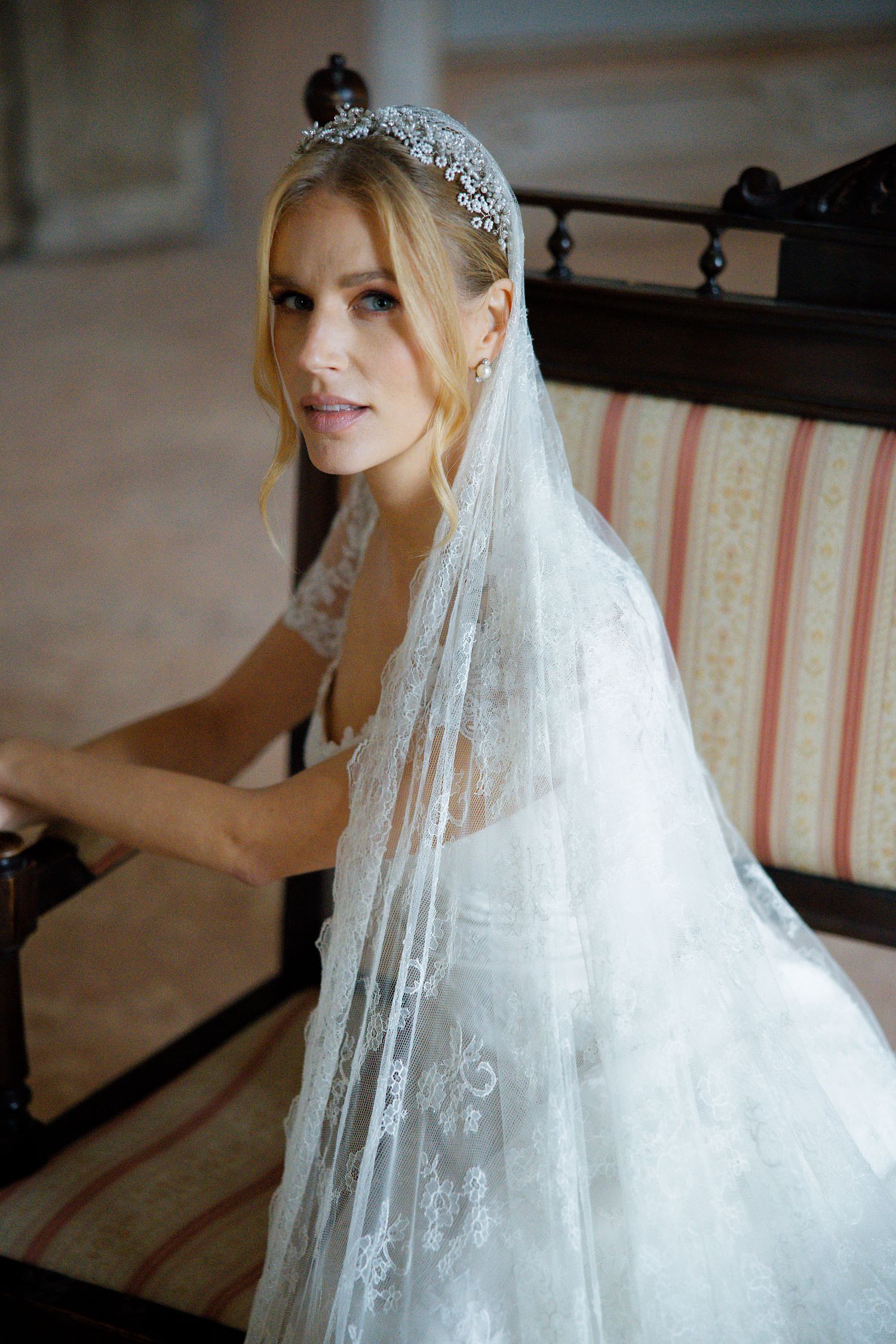 Bride wears delicate lace veil and ornate crystal tiara while seated on striped antique settee.
