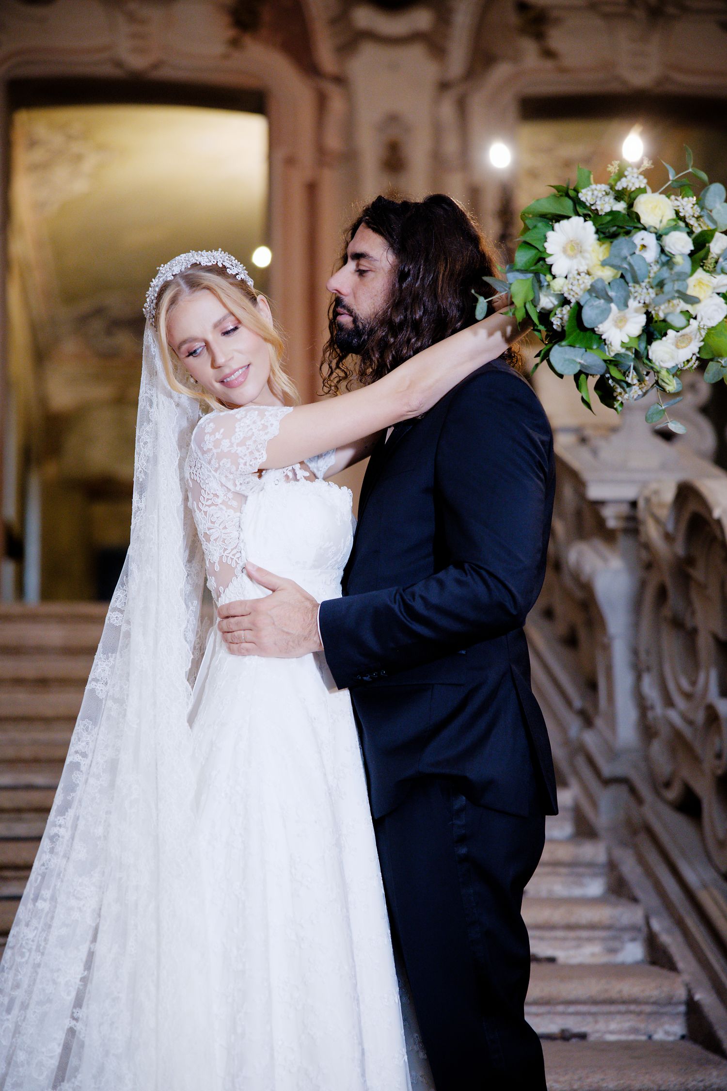 A romantic wedding photo of a couple embracing in an ornate venue with a beautiful white floral bouquet.