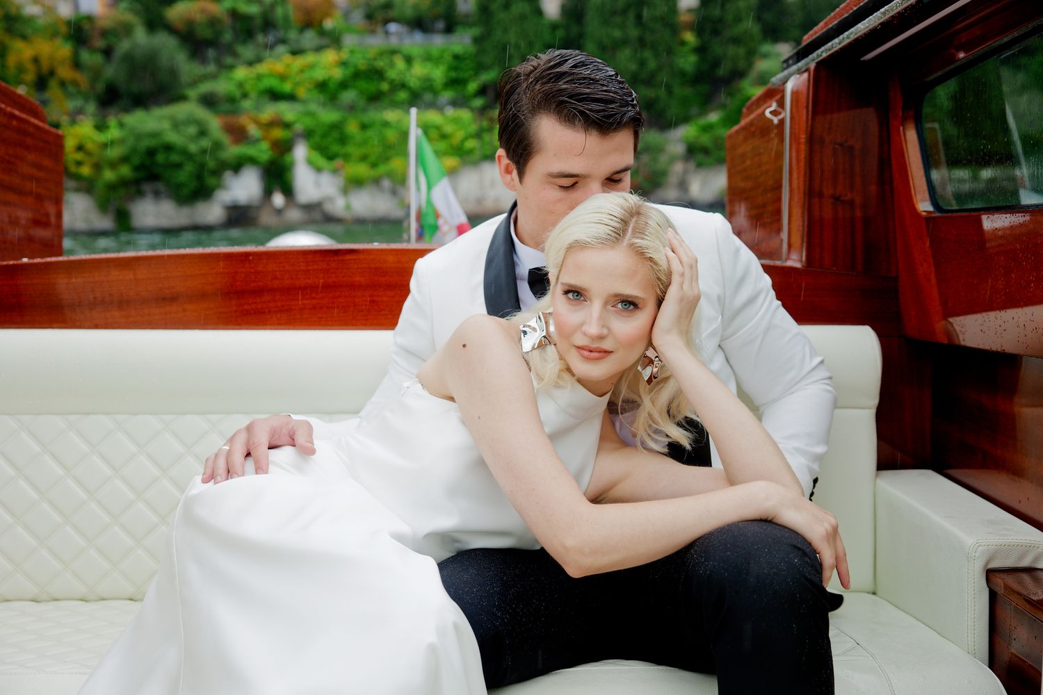 A couple in wedding attire share an intimate moment while resting on white cushions of a wooden boat.