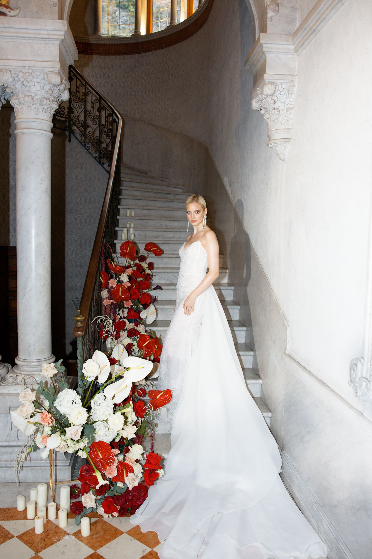Elegant white wedding gown with train next to red and white floral arrangement on grand staircase.