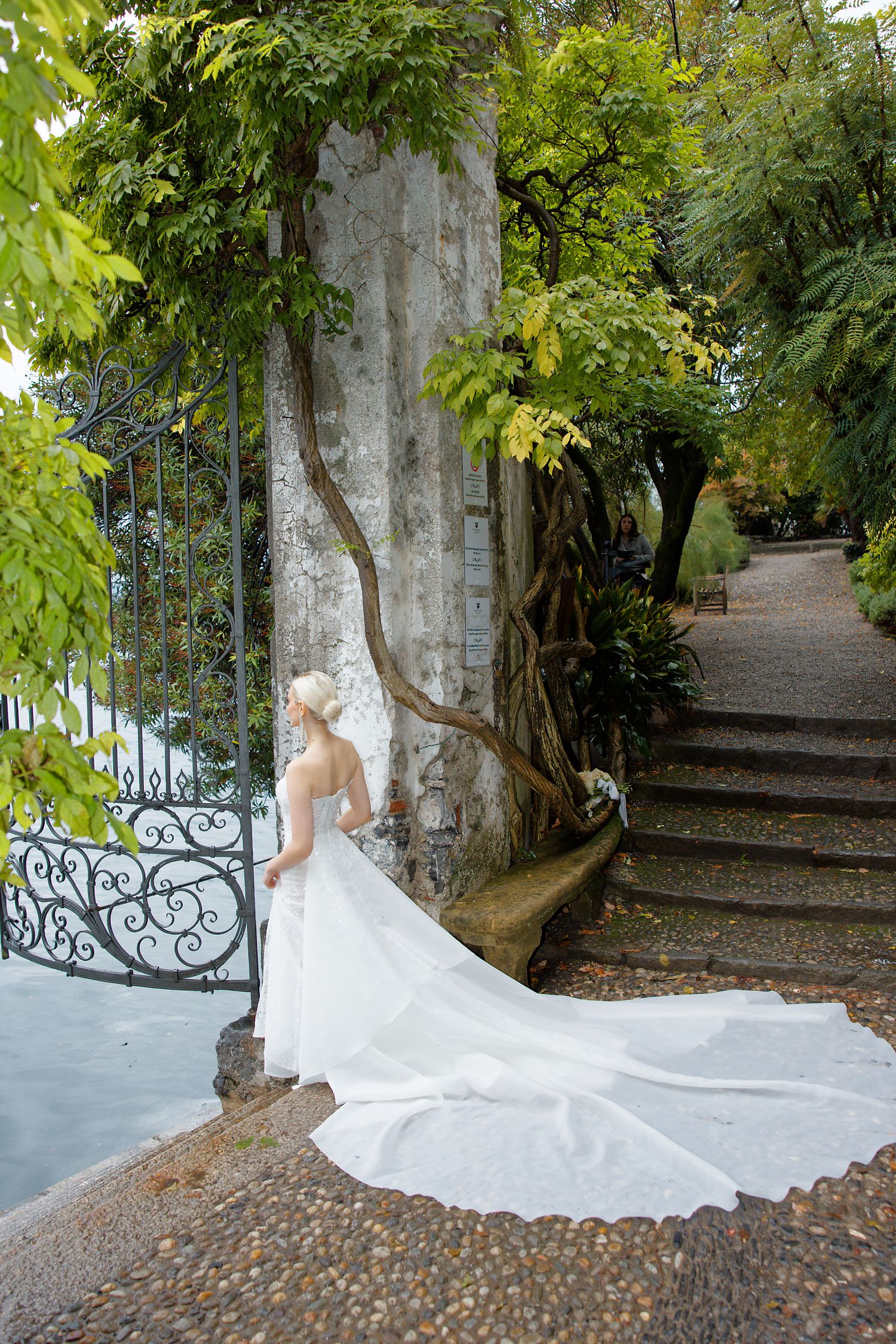 A flowing white wedding dress with long train cascades down stone steps along an ornate iron gate in a lush garden setting.