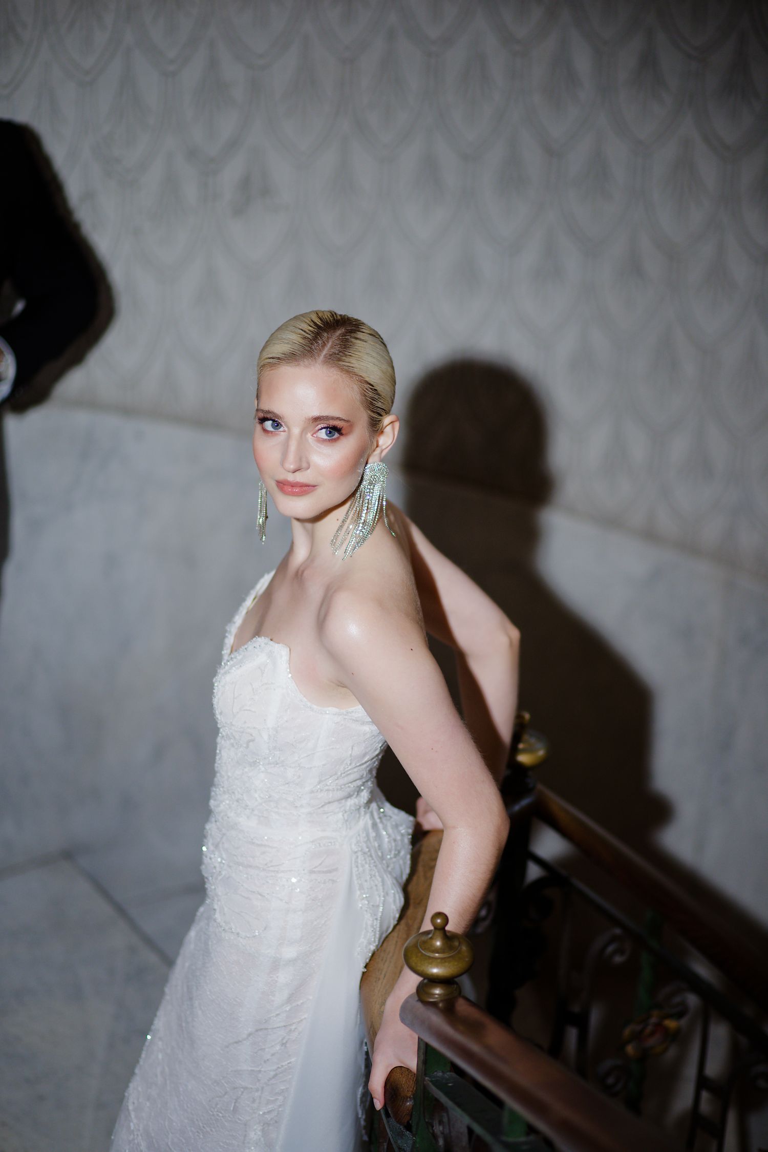Elegant bride in white lace gown poses on staircase with dramatic lighting and wallpaper backdrop.