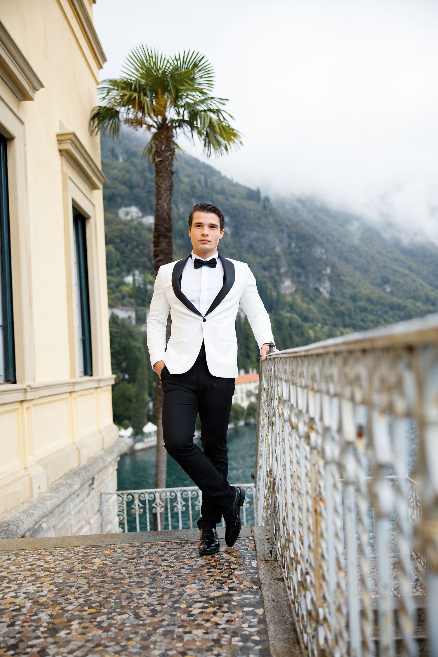 Groom in white dinner jacket stands on ornate balcony overlooking mountain landscape.