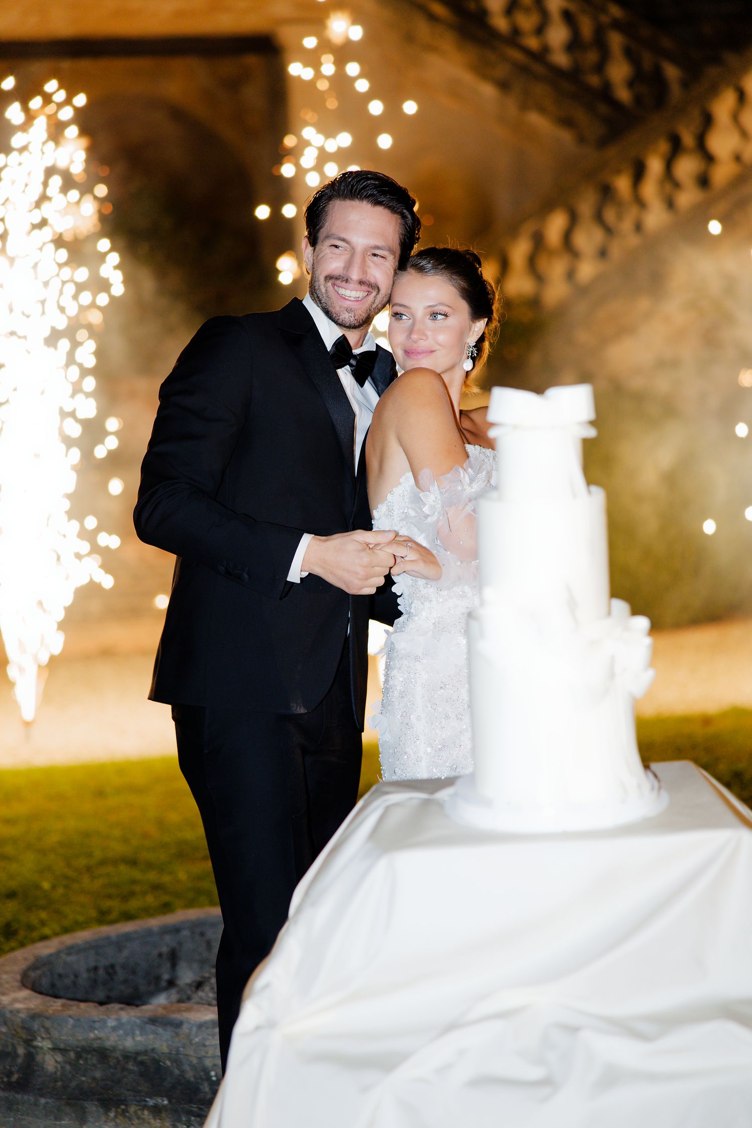 A wedding couple poses with their white tiered cake as sparklers create a magical backdrop.