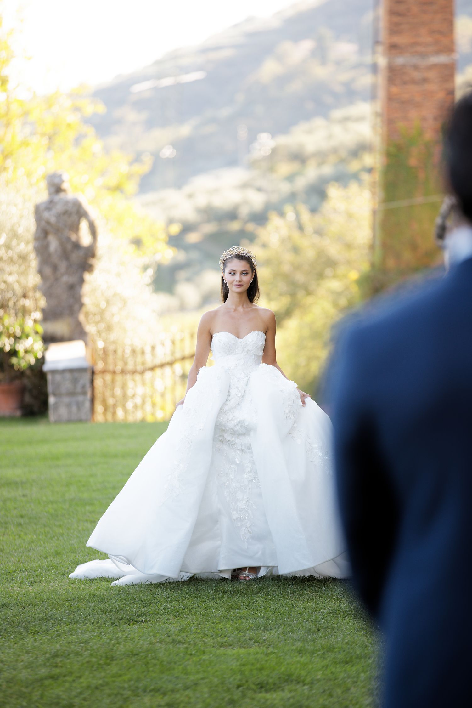 A bride in a flowing ballgown stands on a lawn with mountains and architecture in the background.