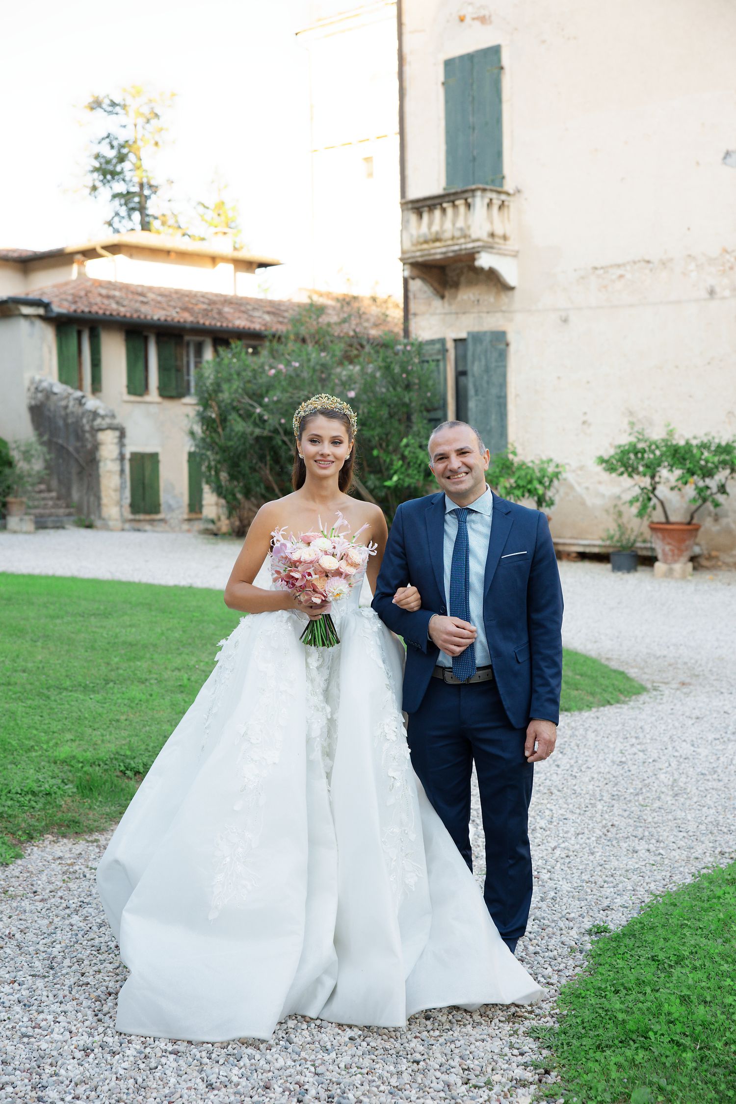 Wedding couple poses together on a gravel pathway near an elegant villa with a manicured lawn and garden.