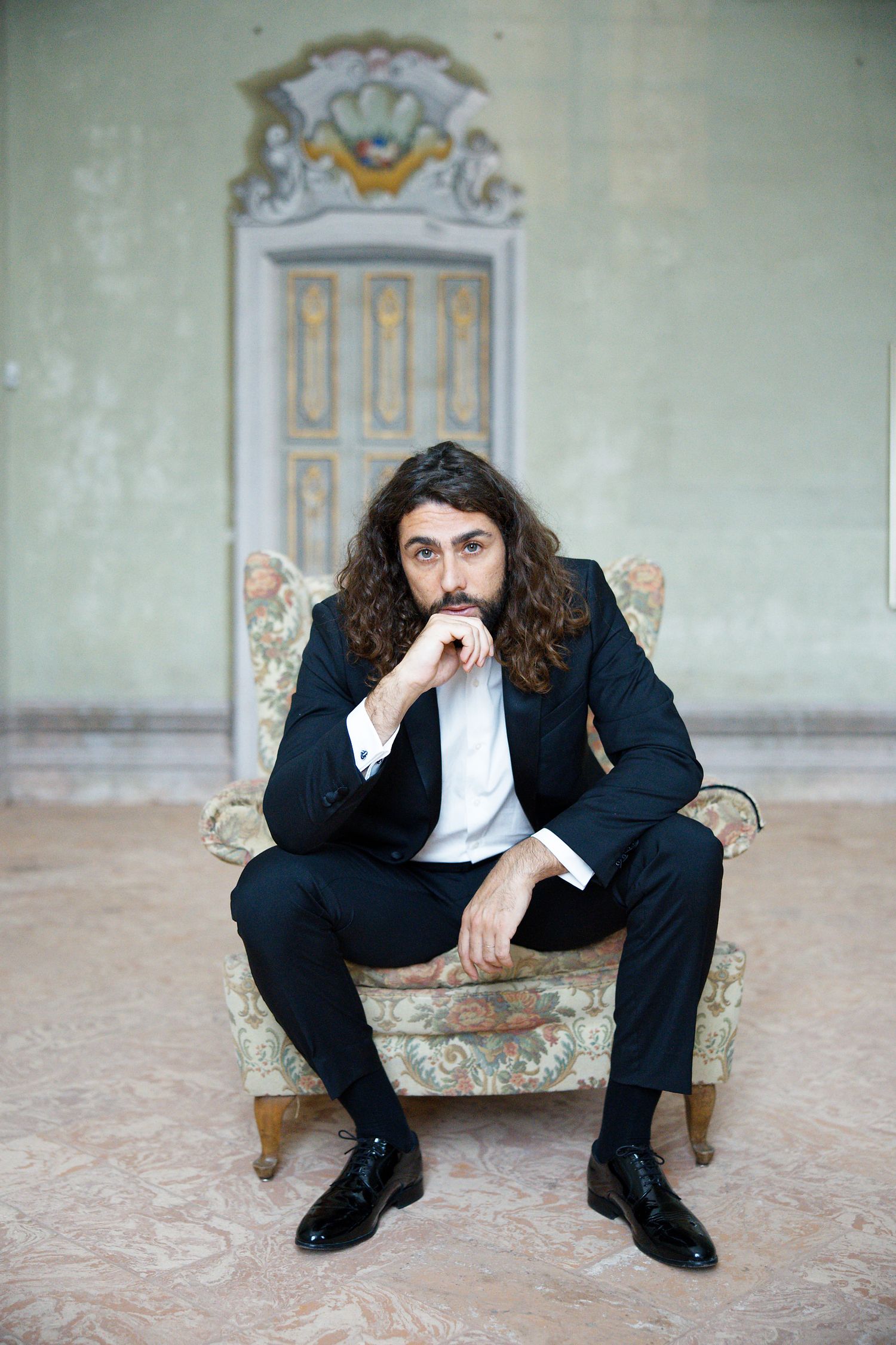 A dark-haired figure in a suit sits thoughtfully in an ornate vintage armchair against a decorative wall with molding.
