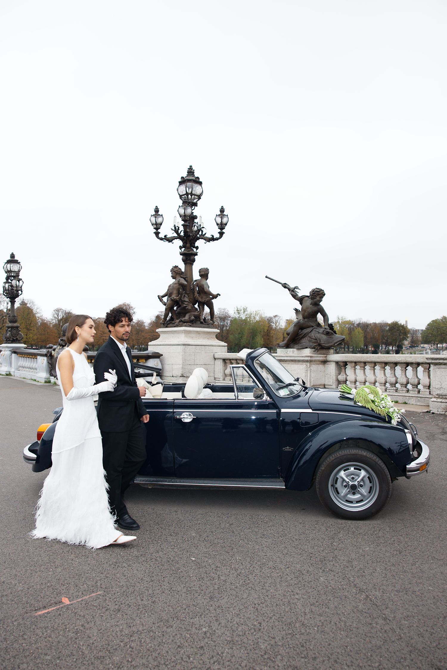 A couple in wedding attire stands beside a classic navy Volkswagen Beetle convertible on an ornate bridge in Paris.