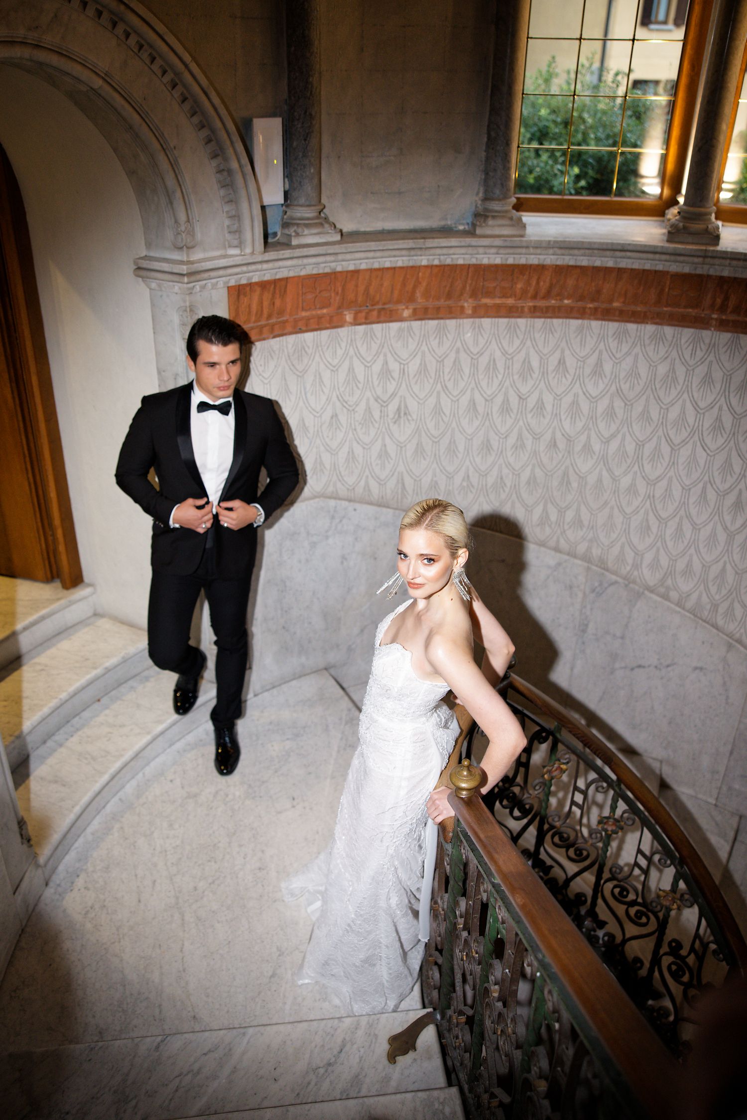 Couple in wedding attire pose dramatically on grand marble staircase.