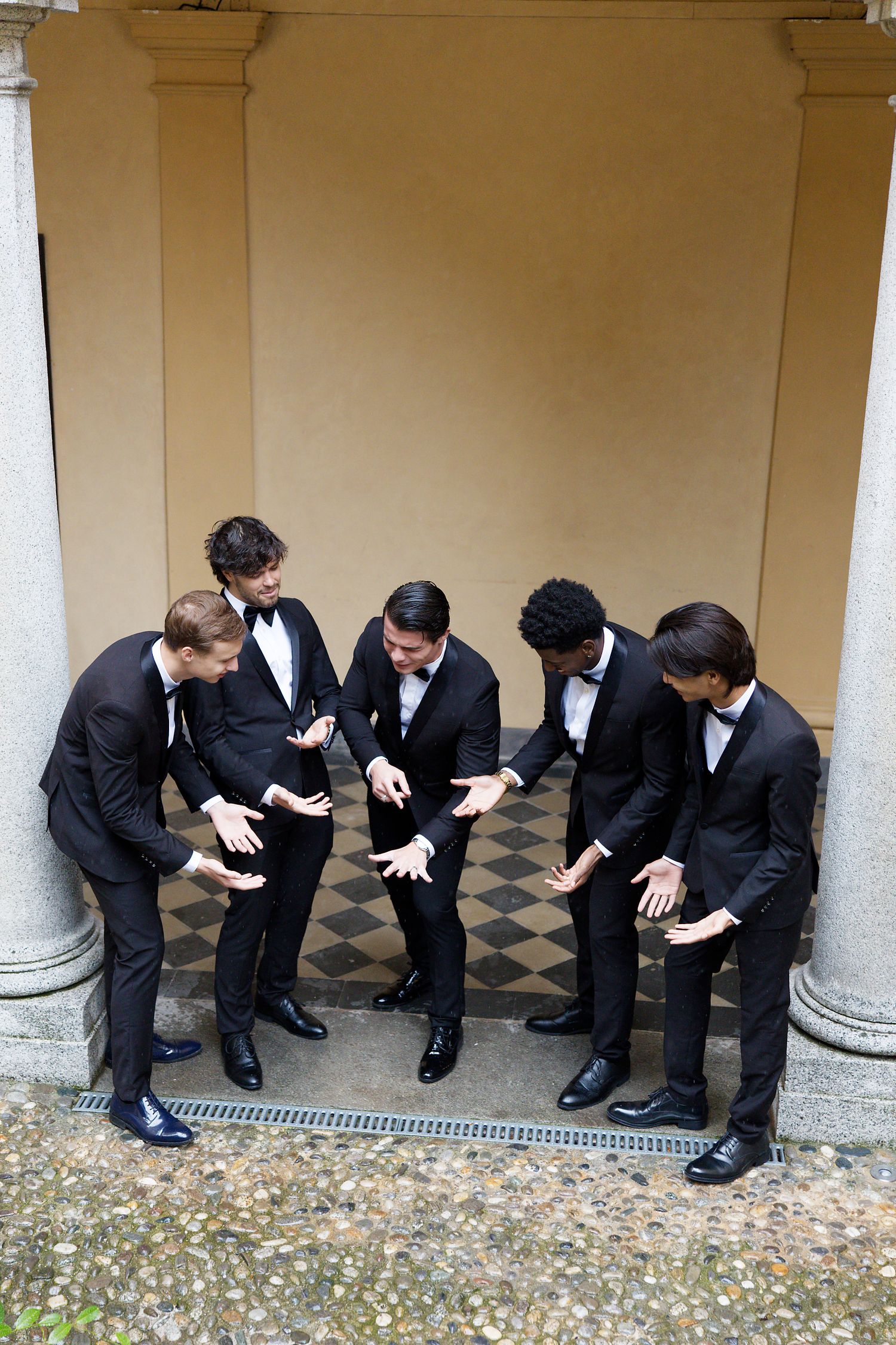 Groomsmen in tuxedos laughing and looking down at their shoes in a formal venue.