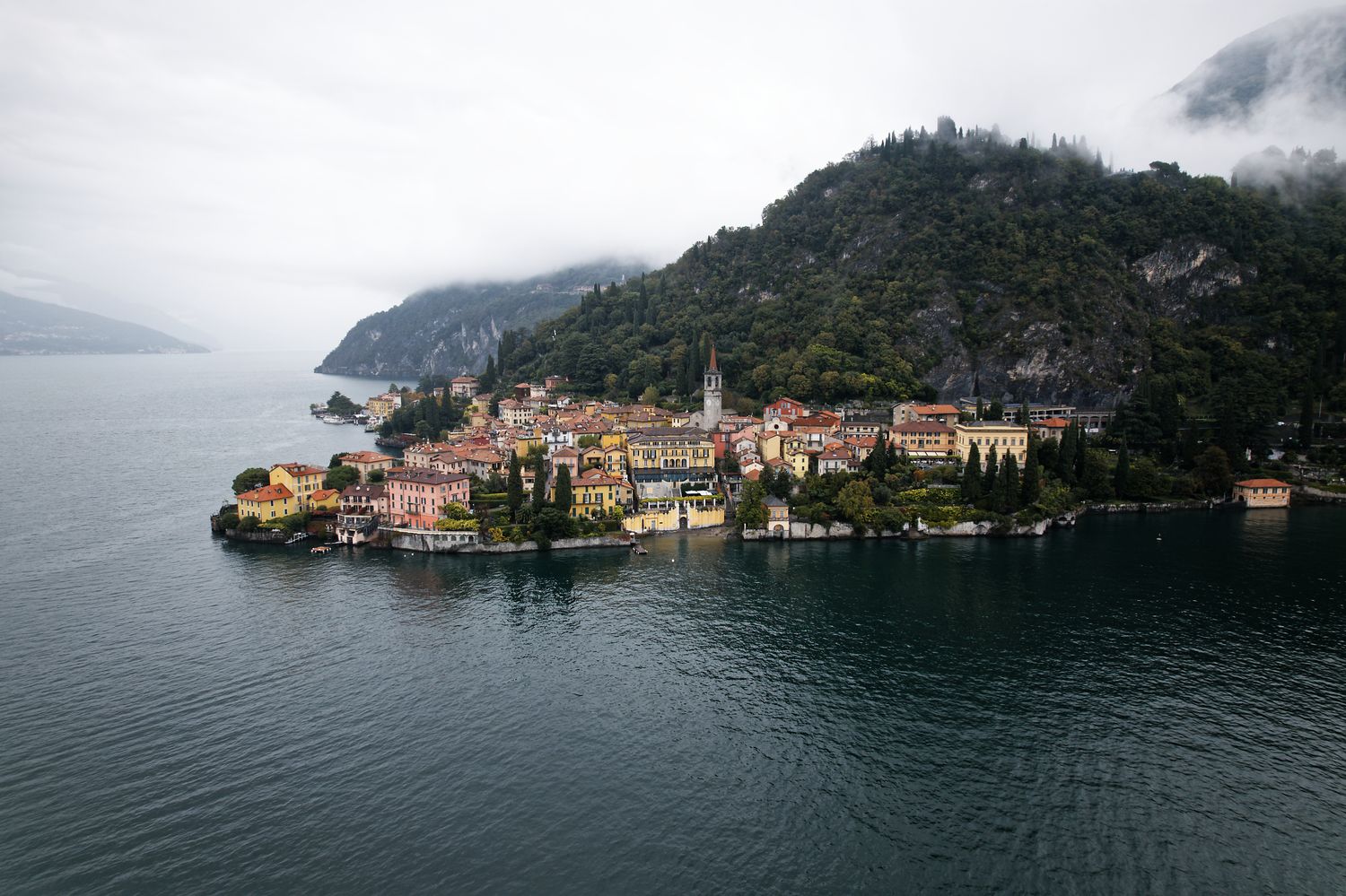 Aerial view of a picturesque Italian village nestled on a peninsula along Lake Como with misty mountains in the background.