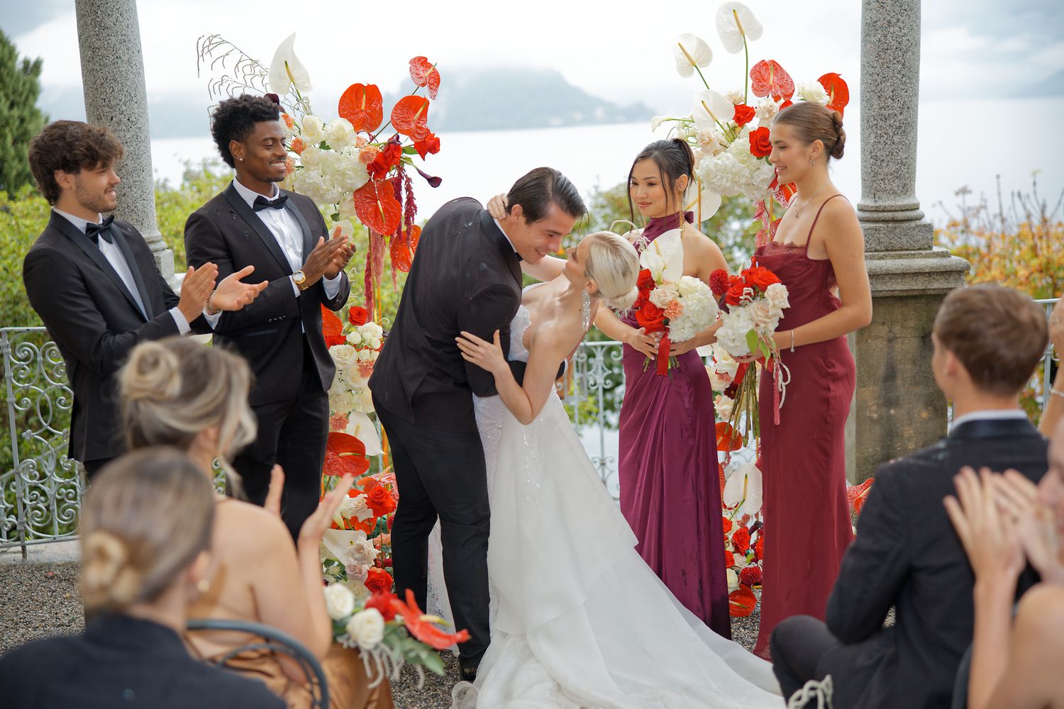 Wedding ceremony guests applaud during a celebration under a pergola with orange floral decorations.