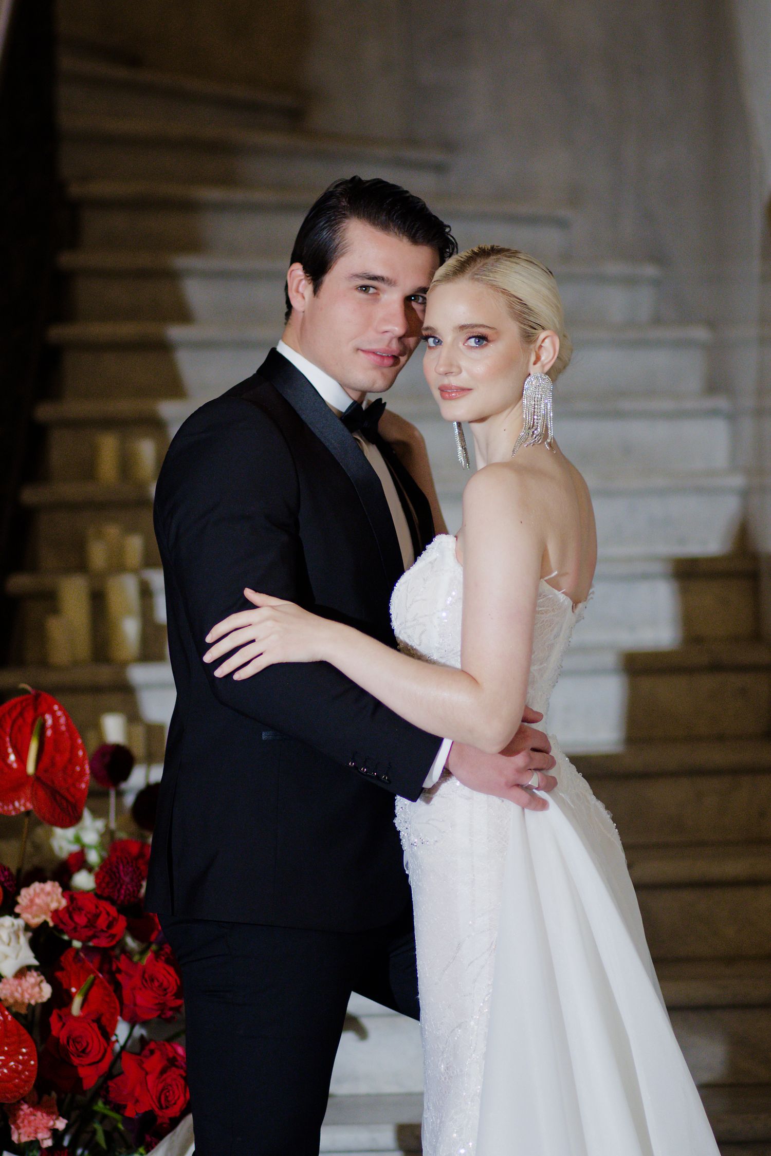A romantic wedding portrait of a couple posing by red roses on a grand staircase.