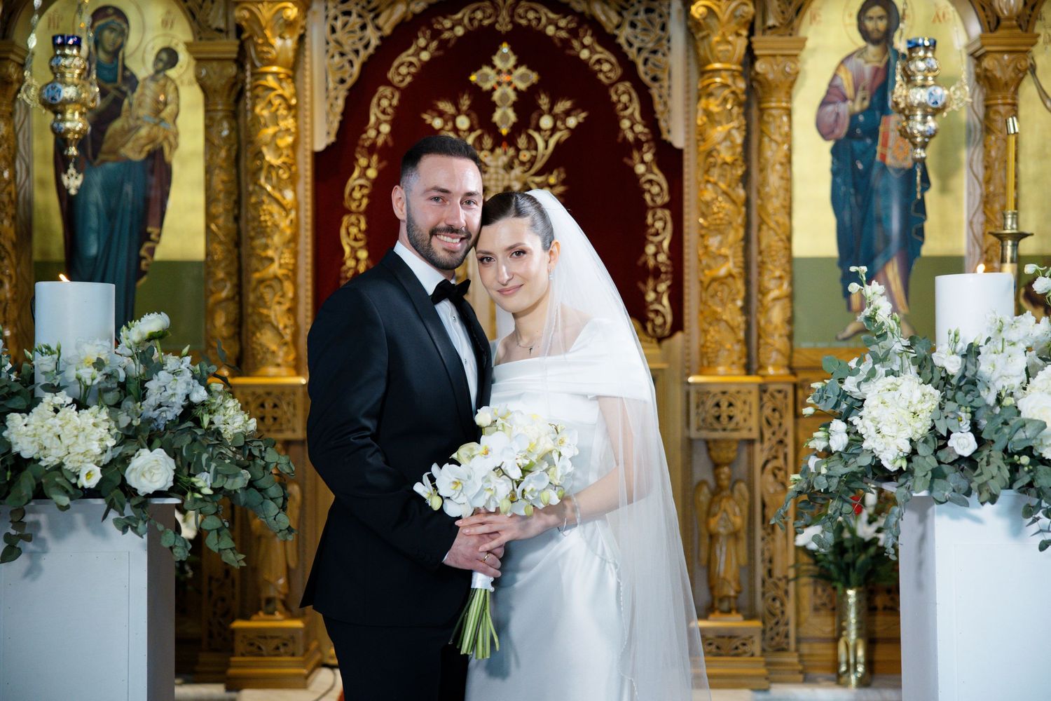 Wedding portrait of couple standing in front of ornate gold altar with white floral arrangements.