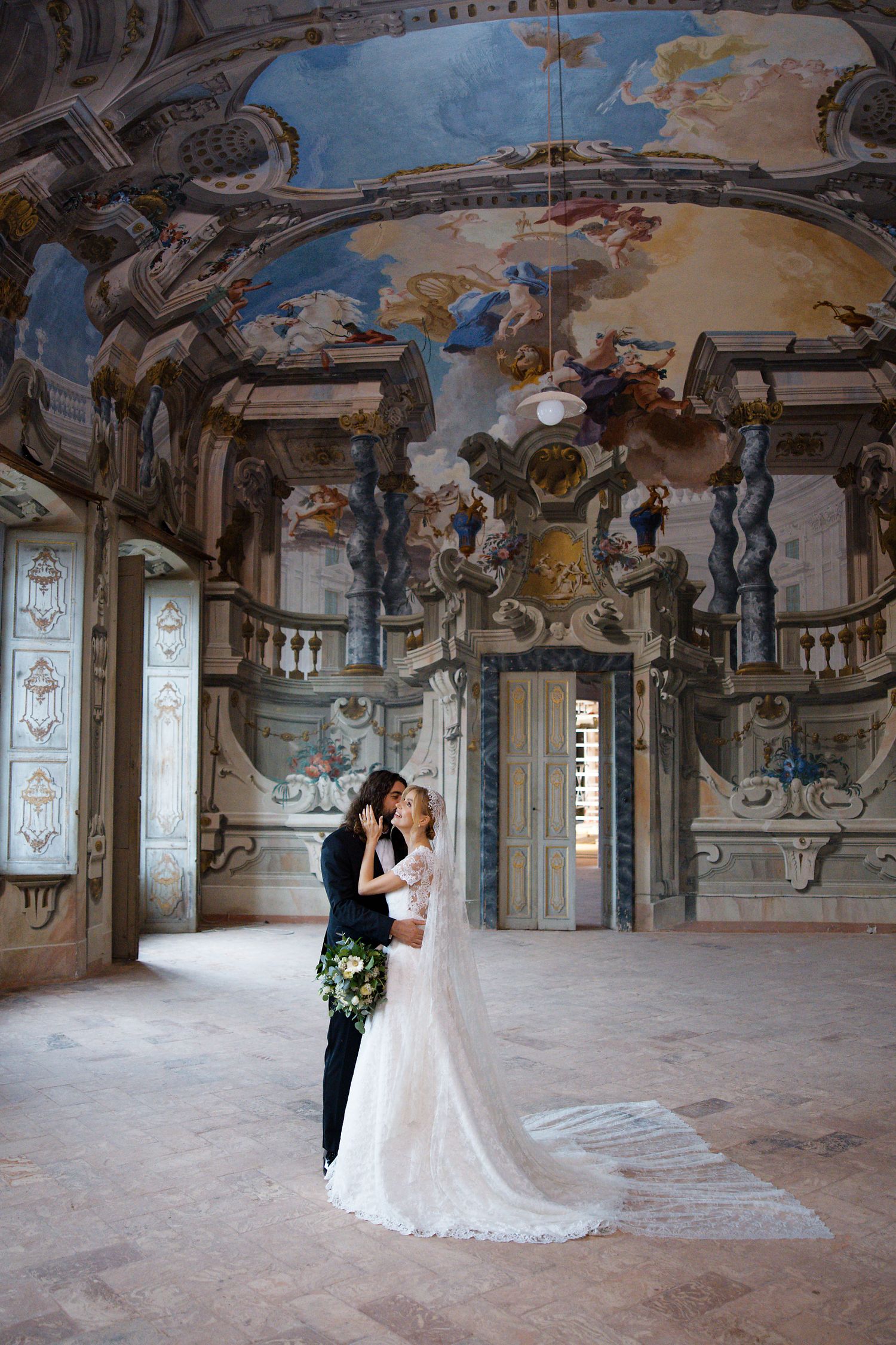 A wedding couple embracing under an elaborately painted baroque ceiling in a historic venue.