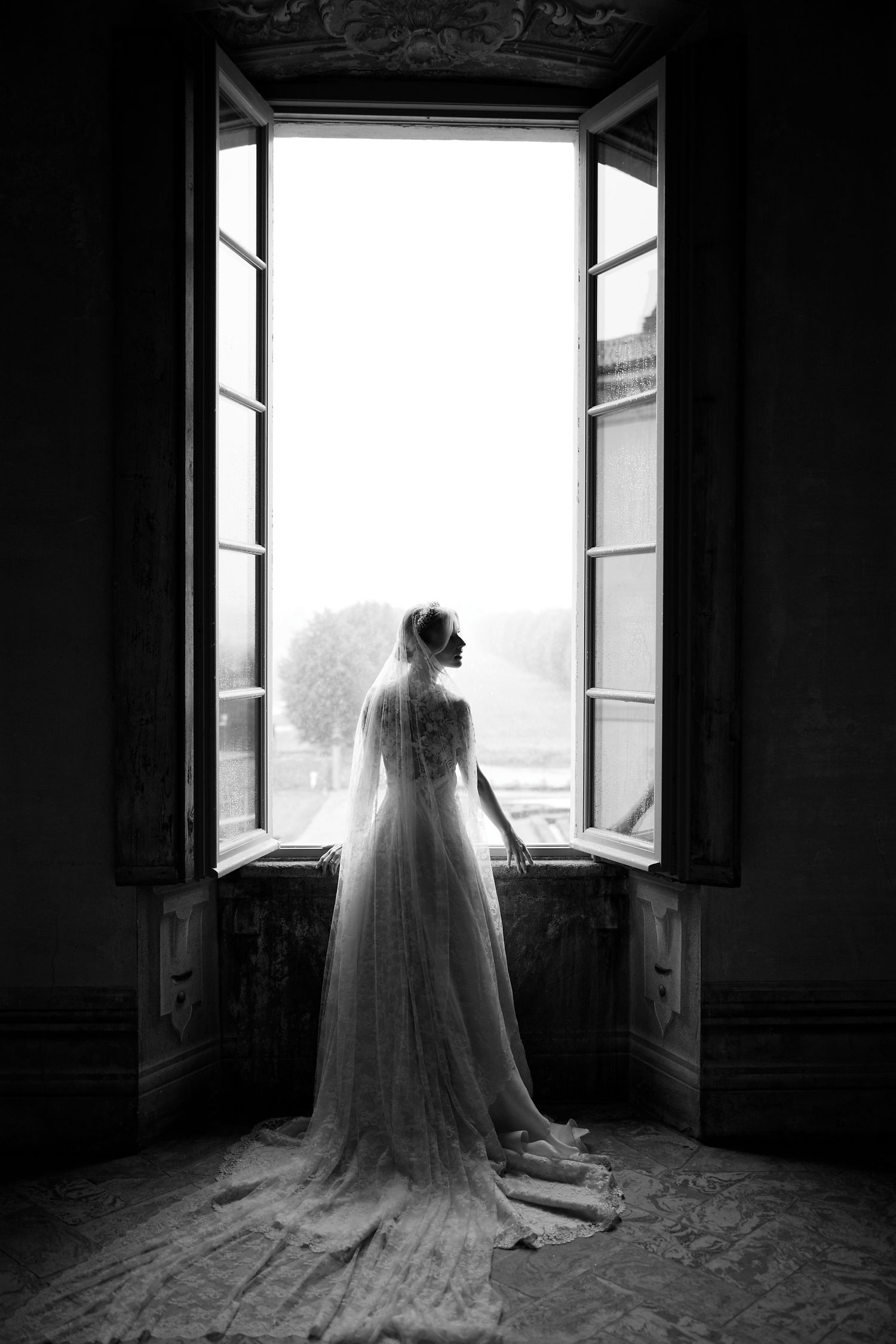 A silhouetted bride in wedding gown stands at an open window in black and white photography.