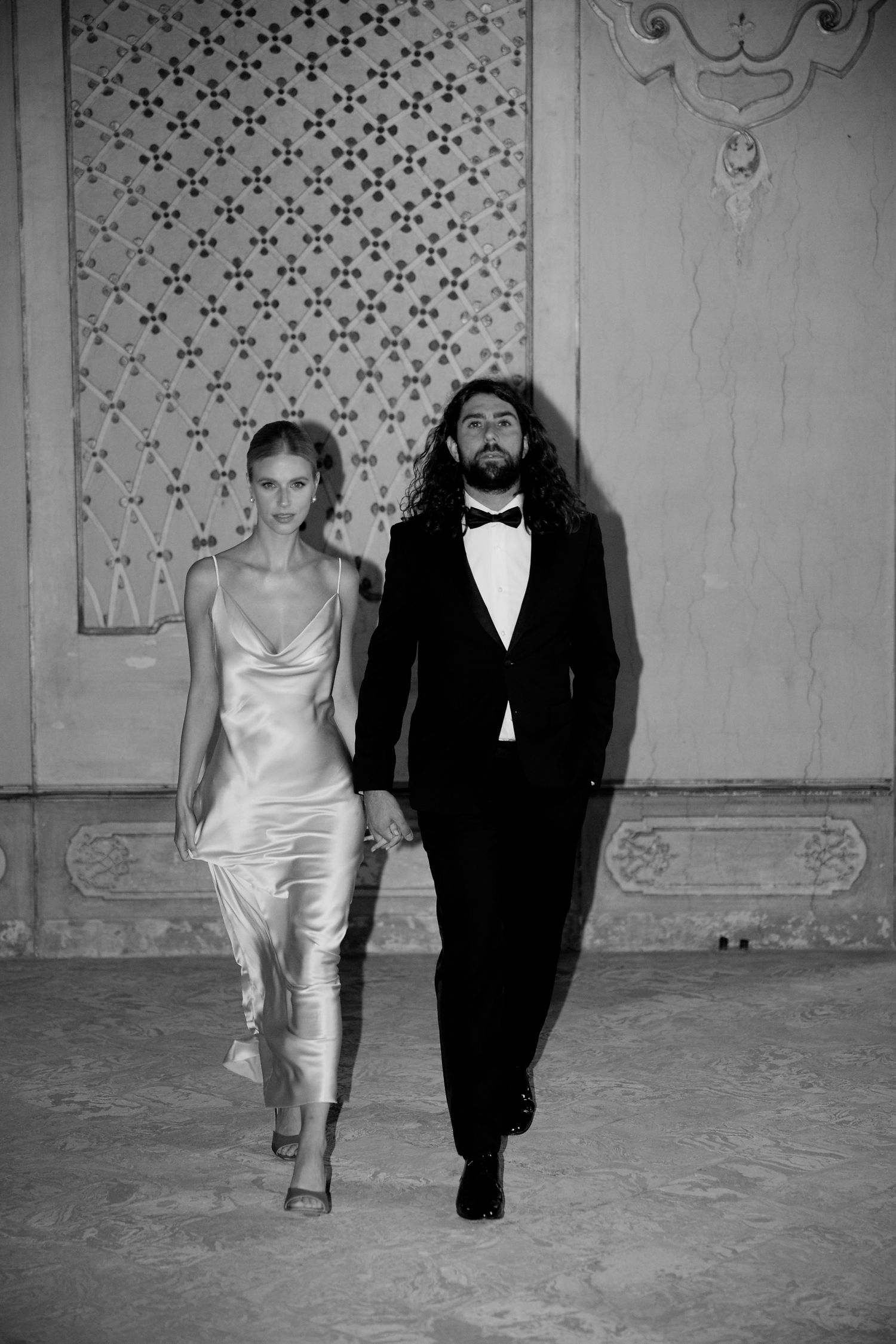 A black and white portrait of a couple in formal attire walking together against a patterned wall.