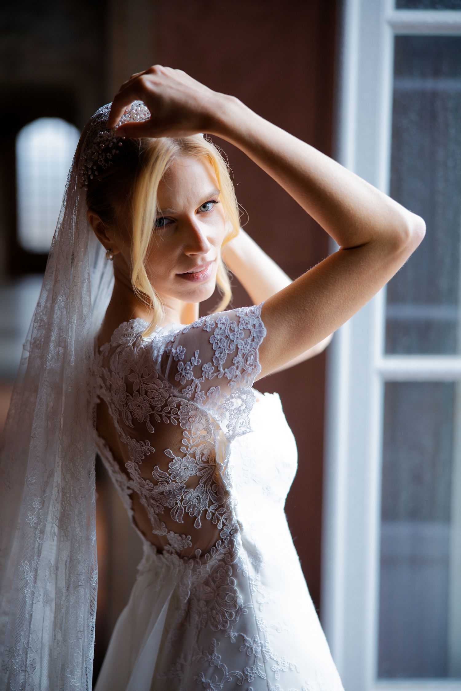 Bride in lace wedding dress poses by window in natural light.