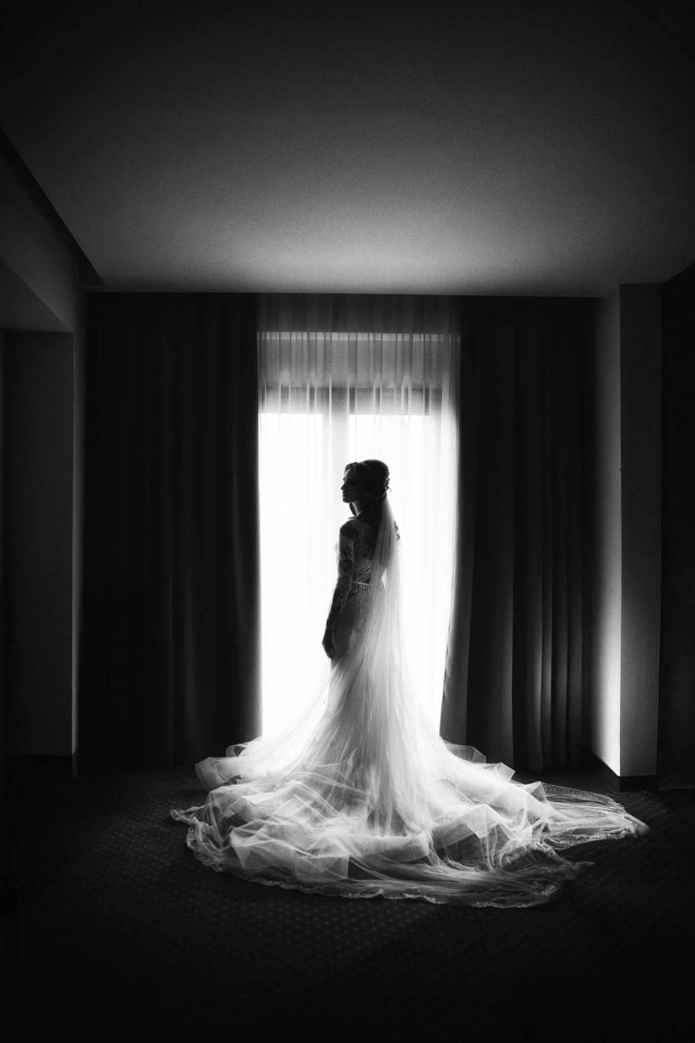 Silhouette of a bride in flowing wedding dress standing against bright window in dark room with dramatic lighting.