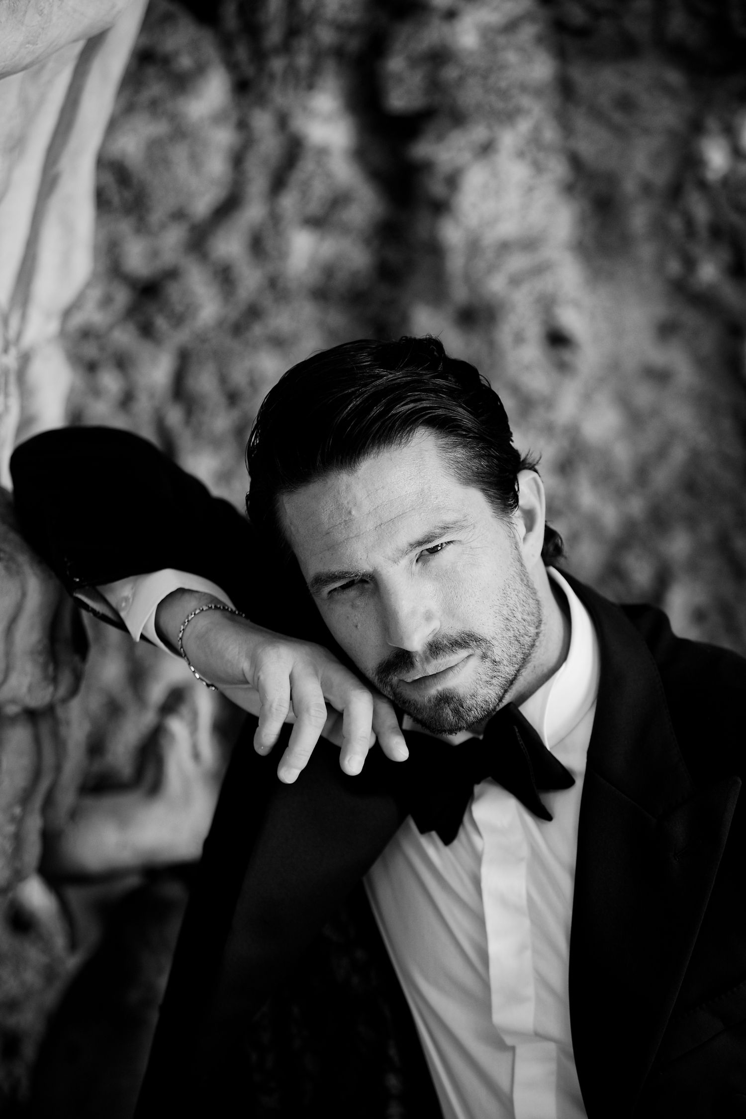 Black and white portrait of dapper gentleman in classic tuxedo and bow tie leaning against stone wall.