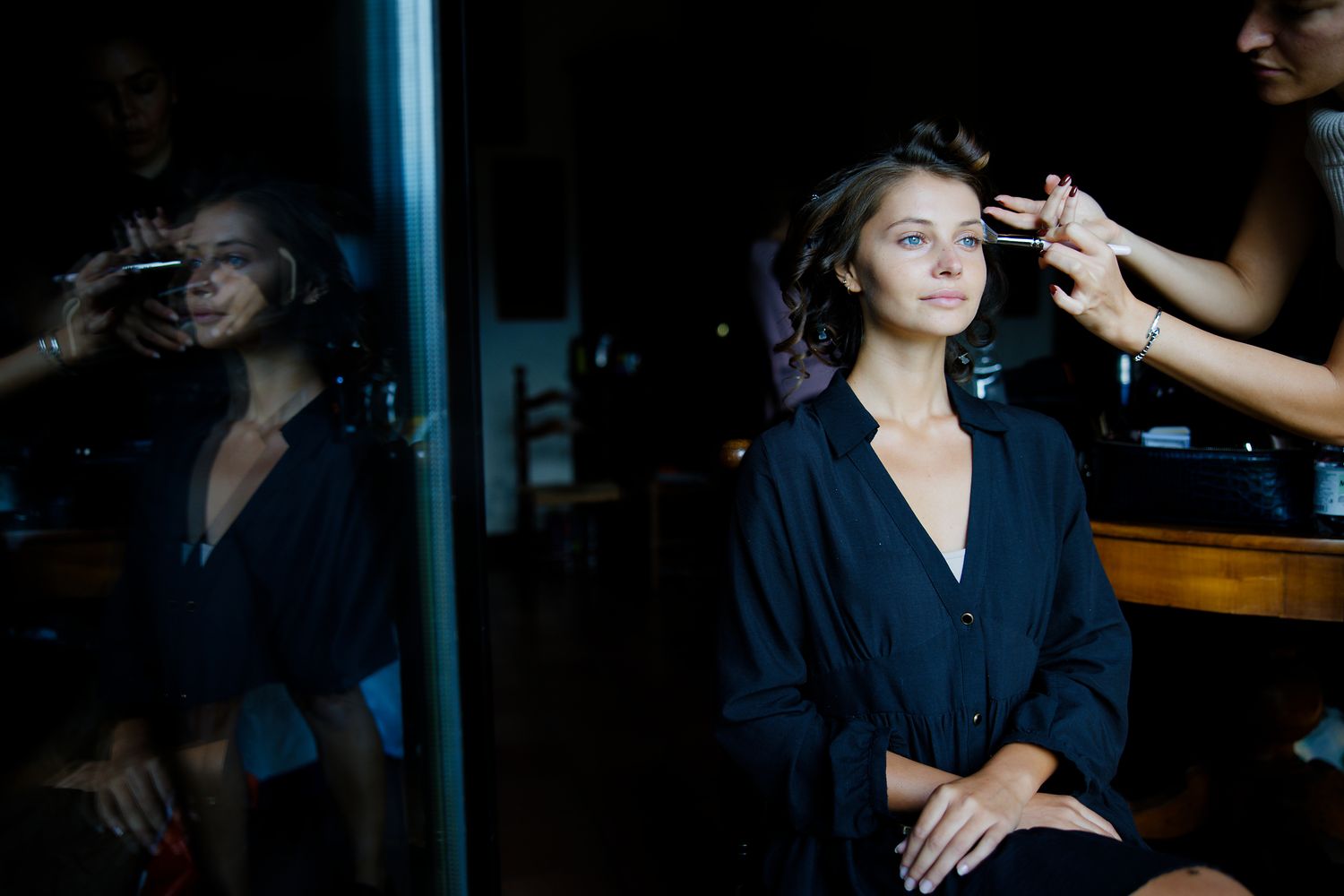 A stylist works on a model wearing a navy blazer in a dark room with mirror reflections.