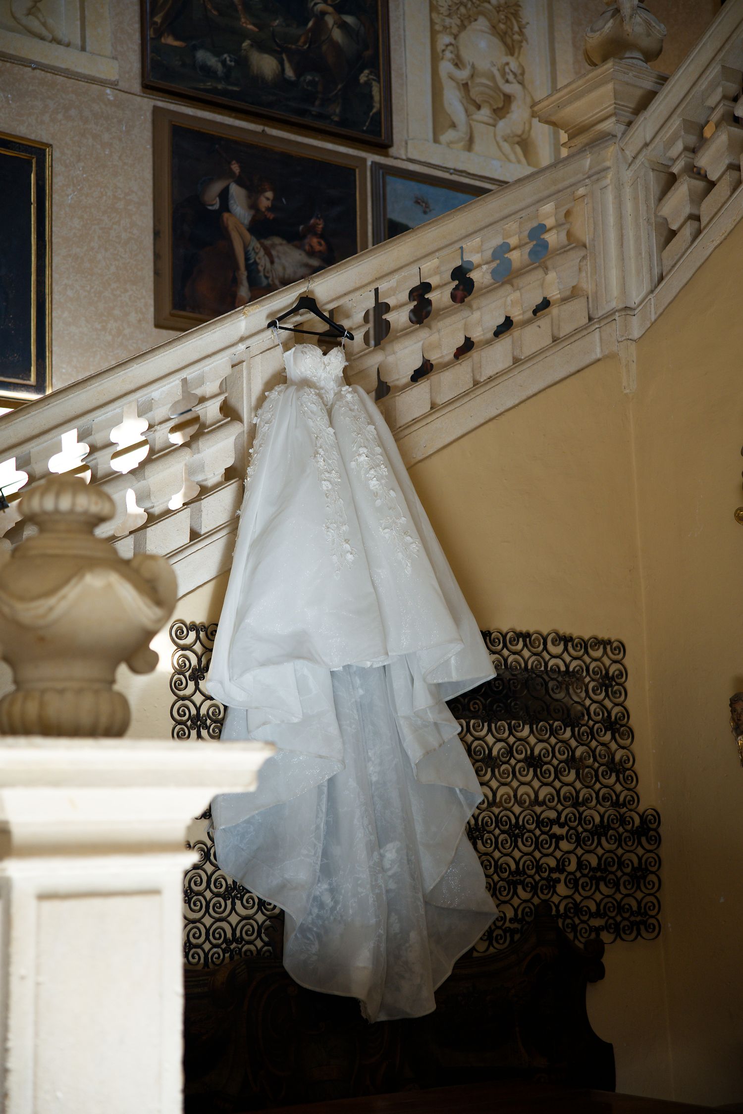 White lace wedding dress hanging on ornate staircase with classic artwork in background.
