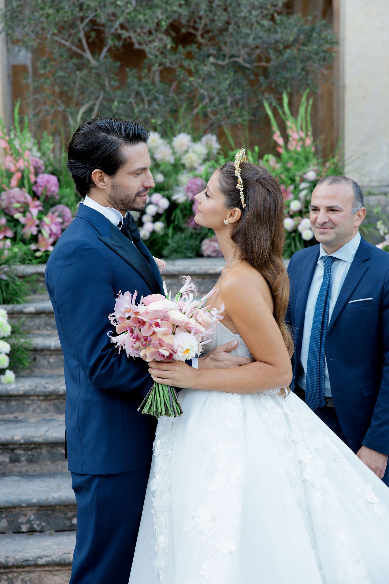 A wedding couple shares a joyful moment on stone steps surrounded by pink and white flowers in a garden setting.