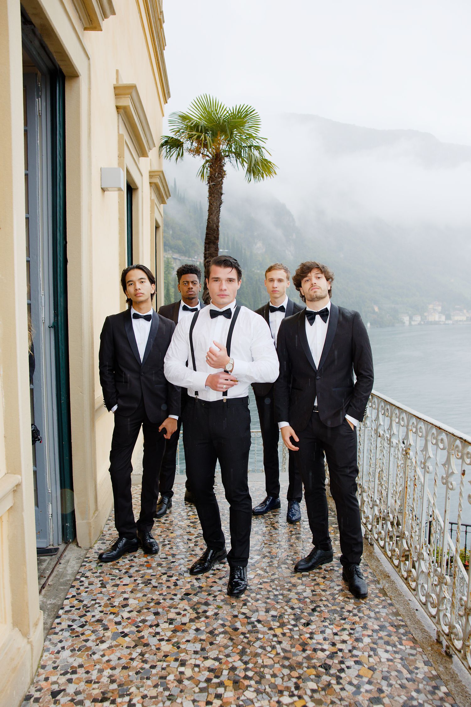 Groomsmen in tuxedos standing on lakeside balcony with mountain views.