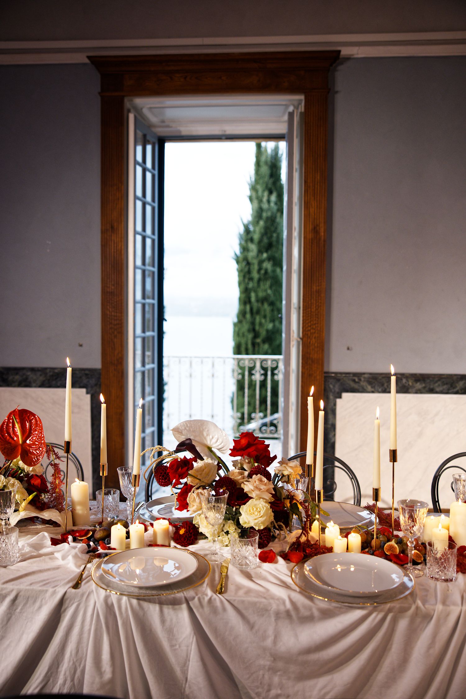 Romantic table setting with candles, red flowers and a scenic balcony view through French doors.