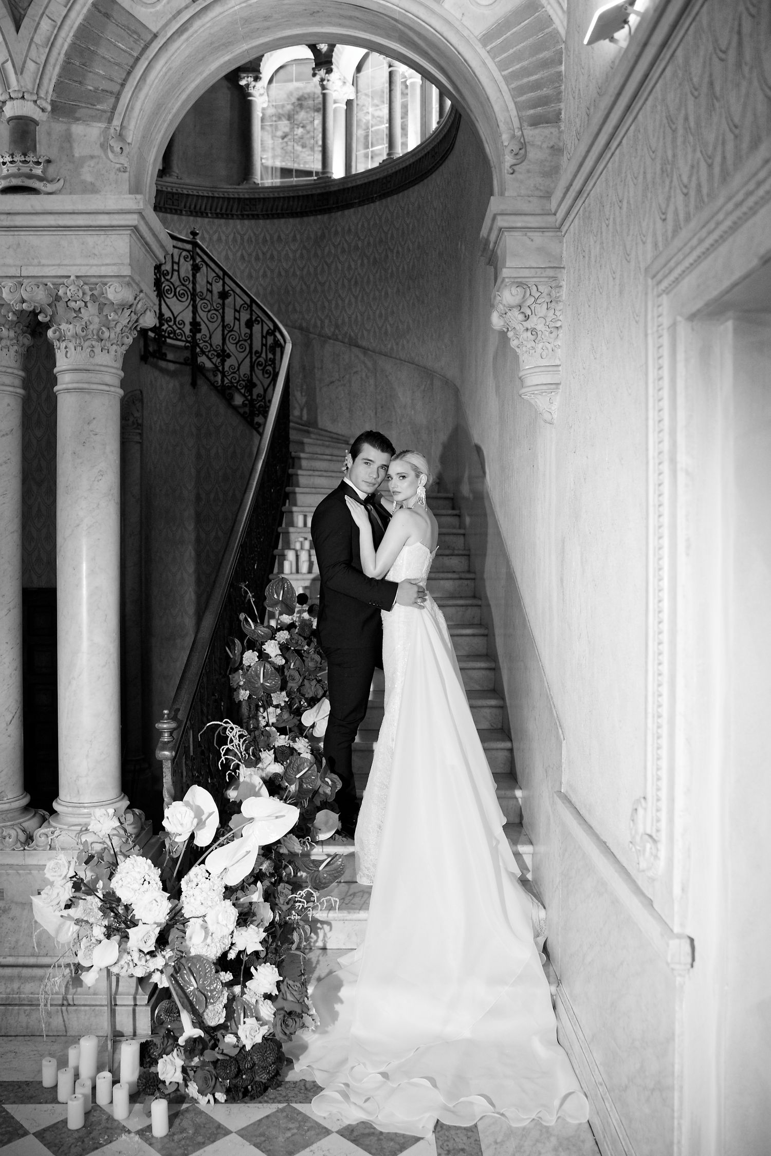 Black and white photo of a romantic wedding moment on an ornate marble staircase with floral arrangements.