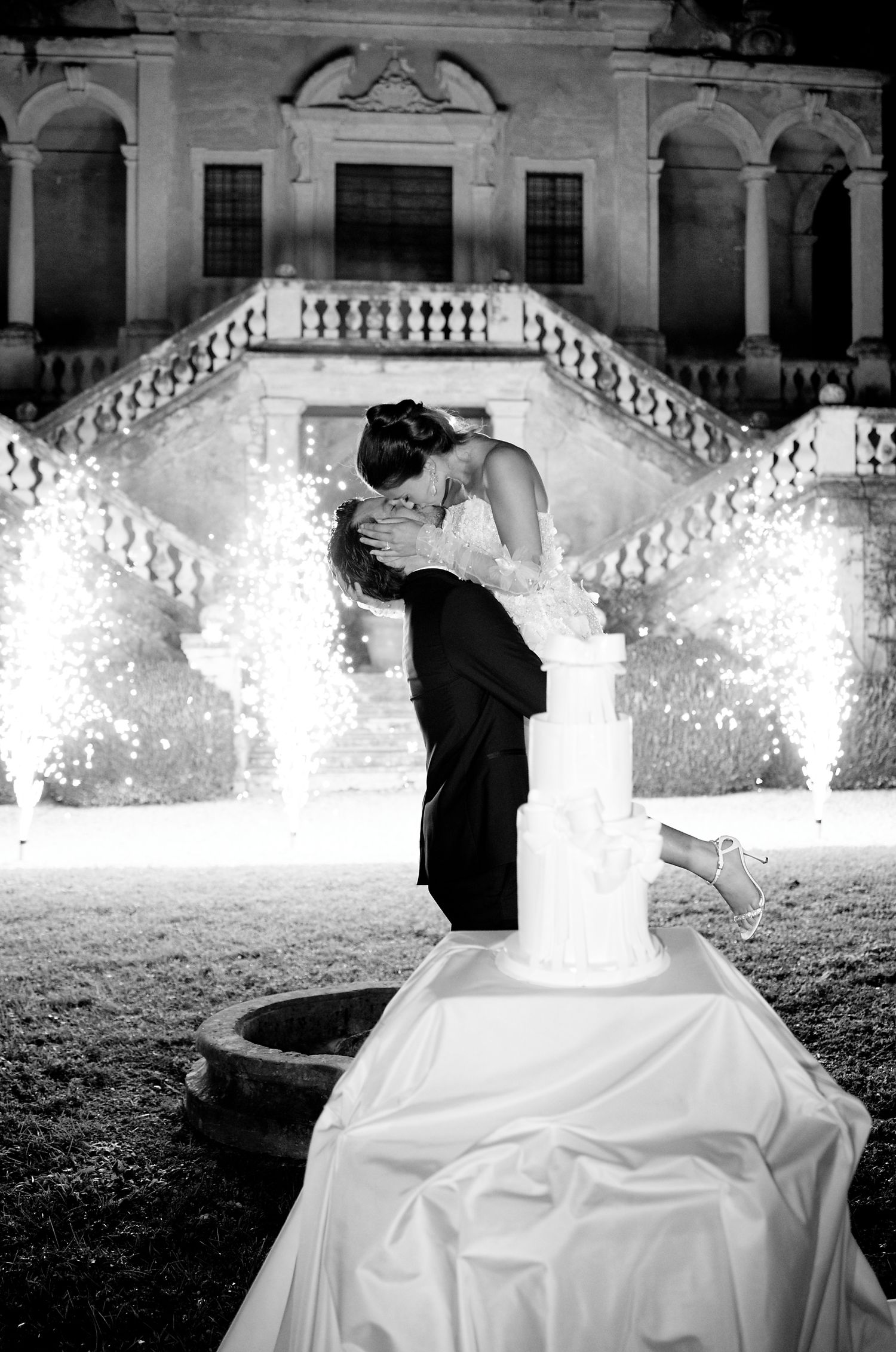 Dramatic black and white wedding photo with couple kissing by their cake as sparklers illuminate the scene.
