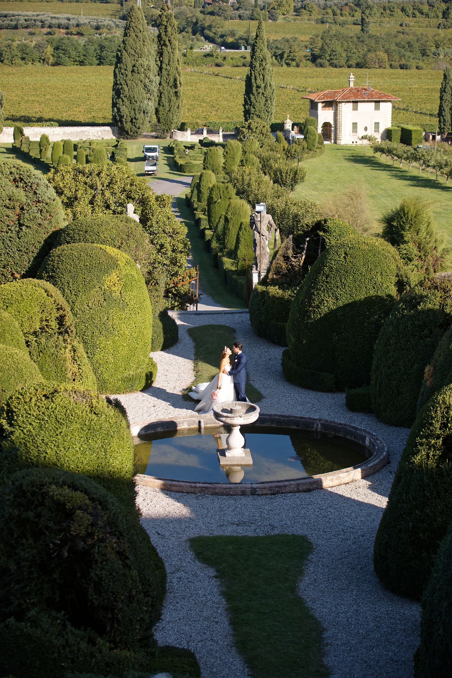 A formal garden features manicured hedges and a decorative fountain where people gather at a luxury Italian villa.