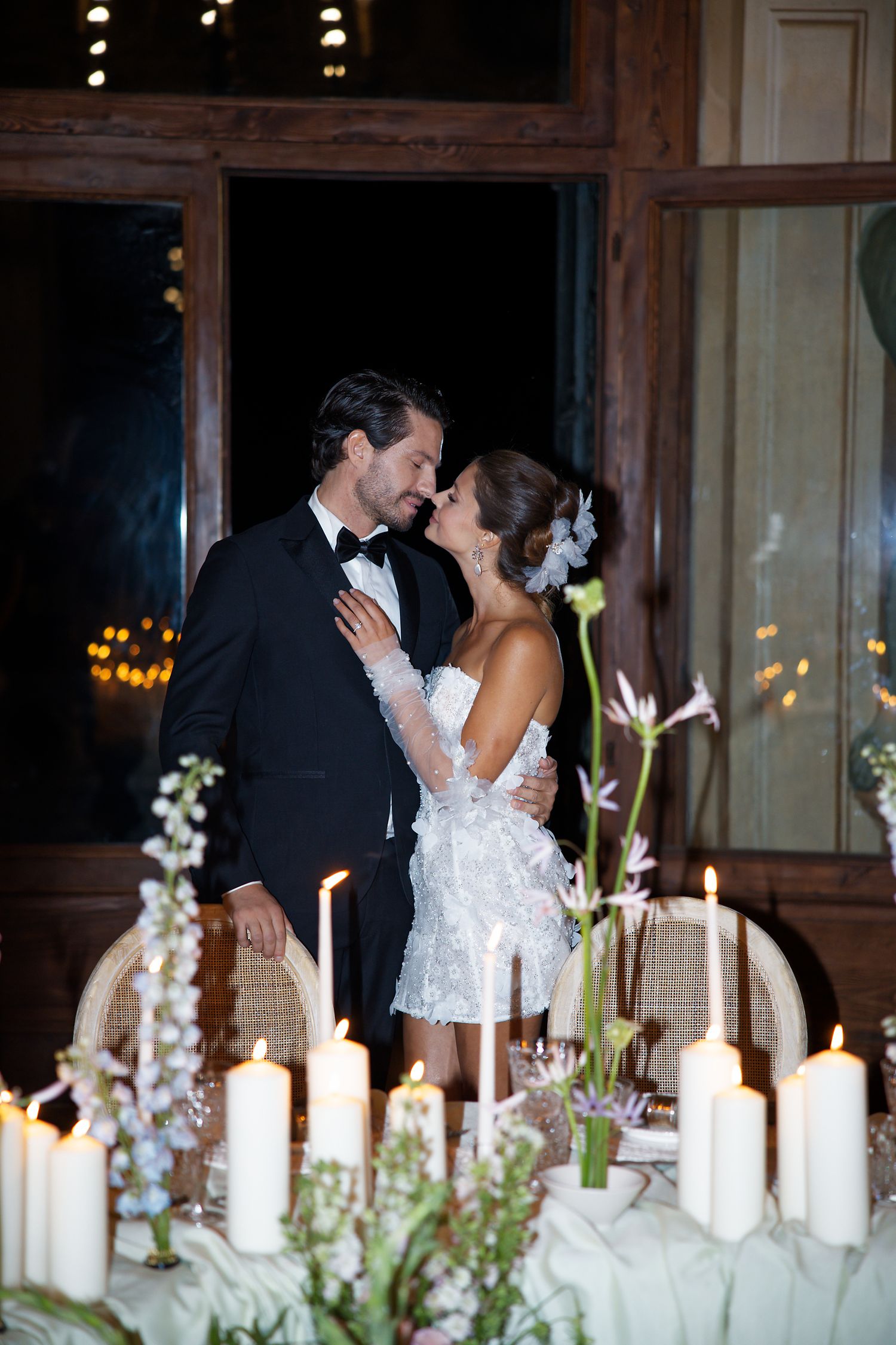 Romantic couple embraces by candlelit table with floral arrangements.