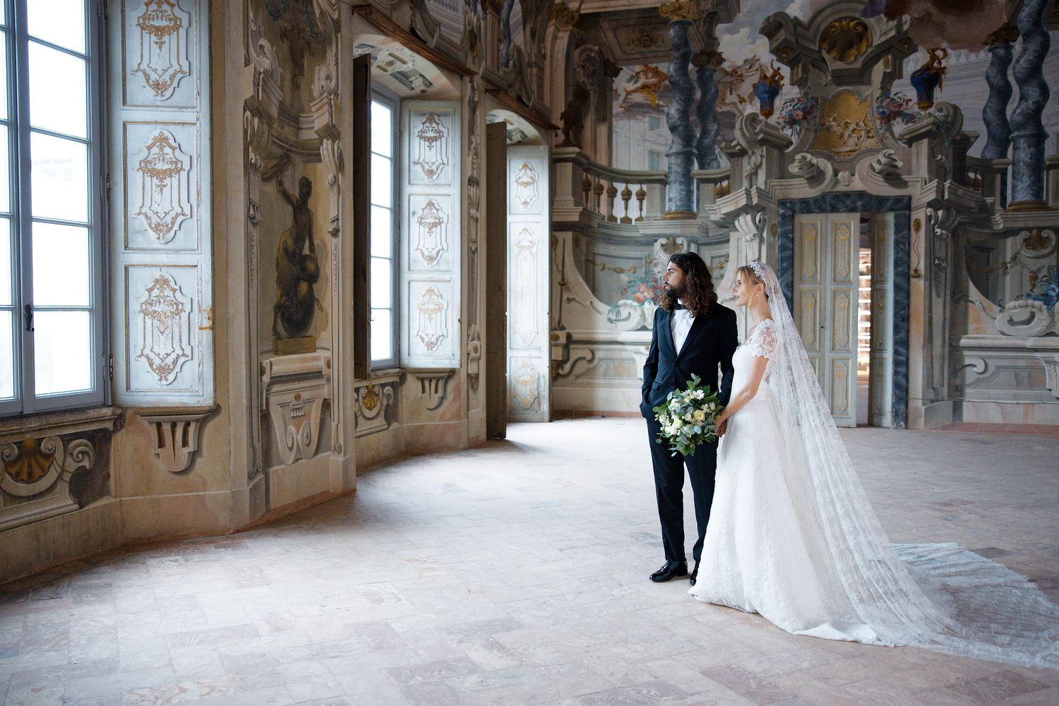 An elegant wedding couple stands in a historic palace room with ornate wall decorations and large windows.