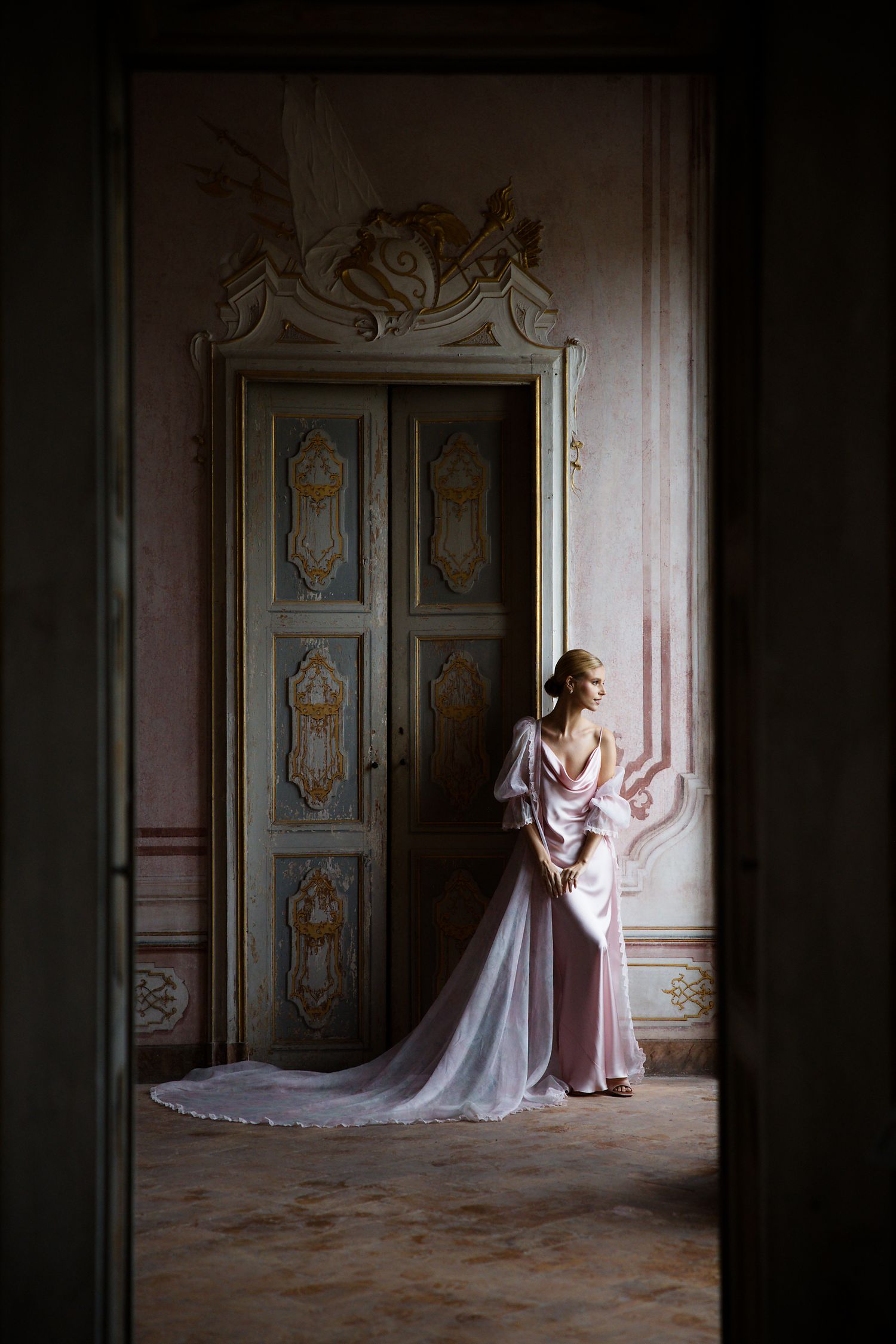 A bride in flowing pink gown stands dramatically against ornate vintage doors in a historic space.