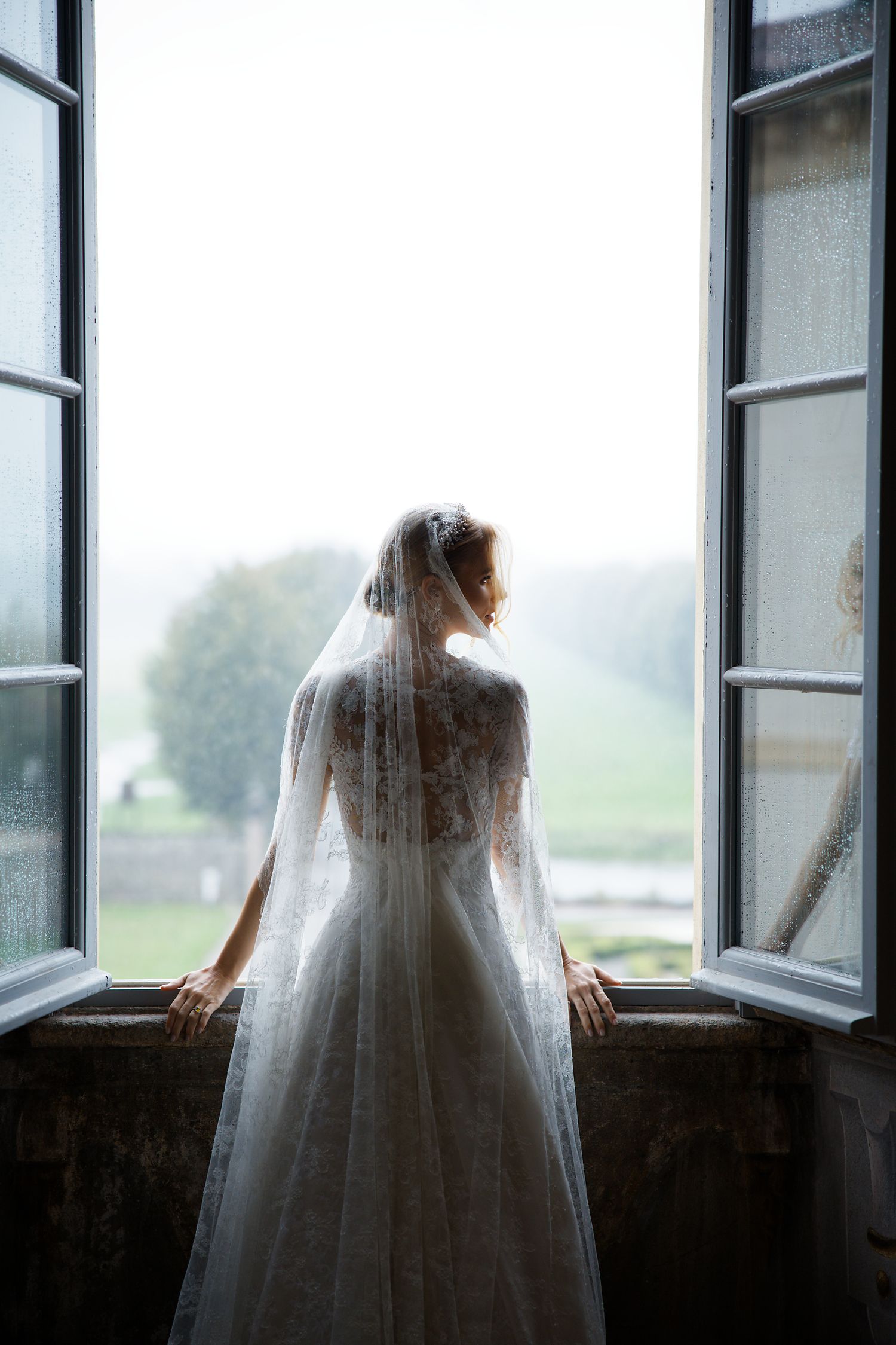 Bride in lace dress silhouetted against bright window overlooking countryside.
