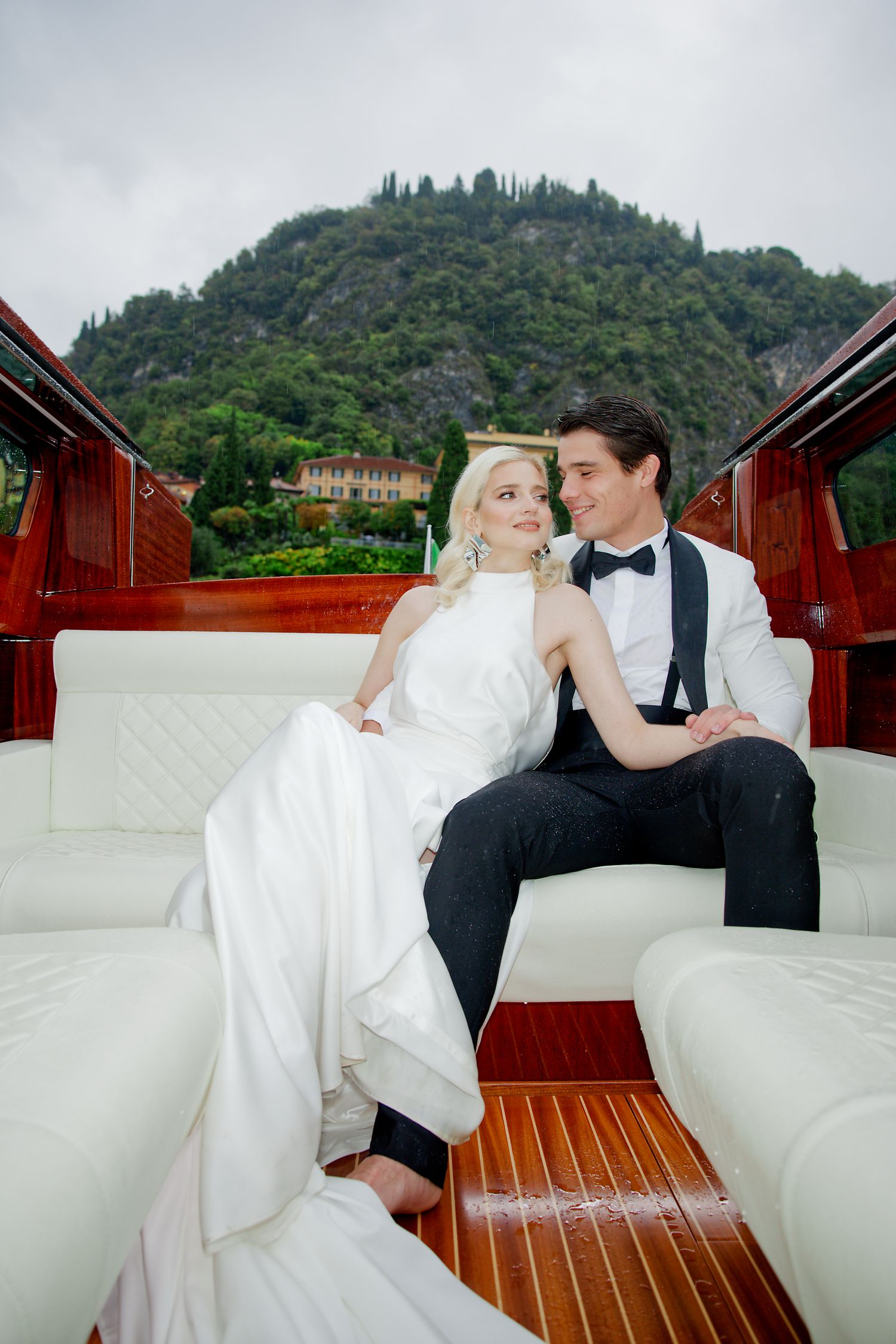 A wedding couple relaxes on white cushions in a wooden boat with Italian hillside villas in the background.