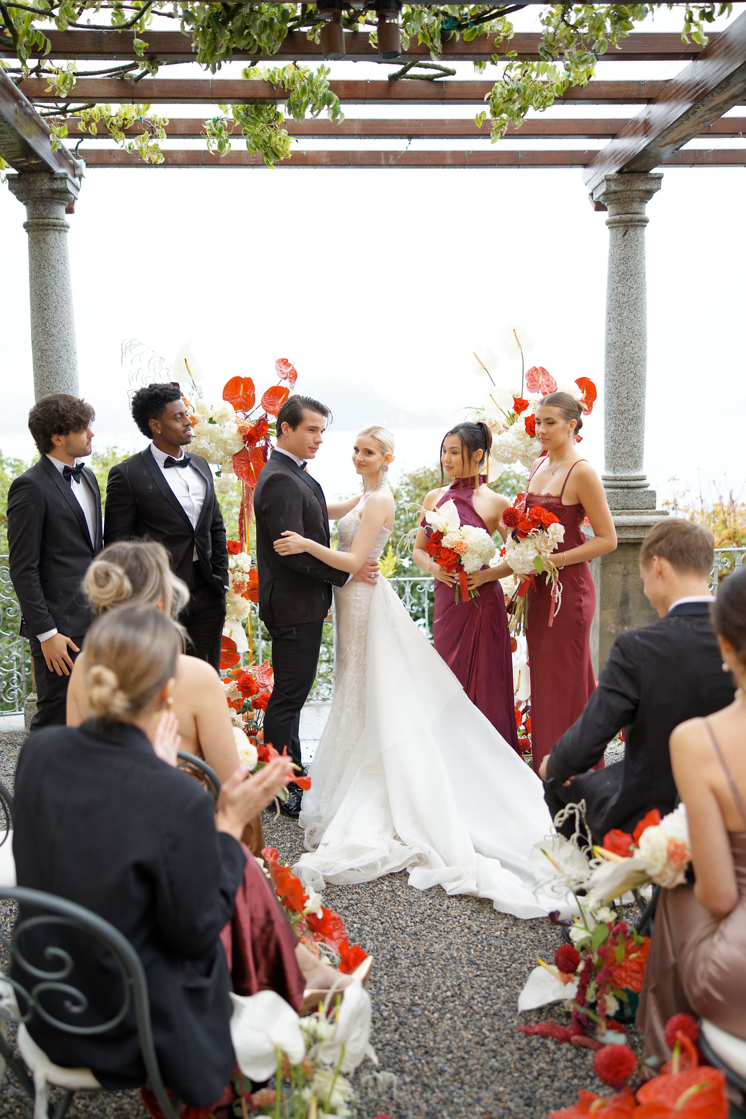Wedding party gathered under a vine-covered pergola with burgundy and white floral decorations.