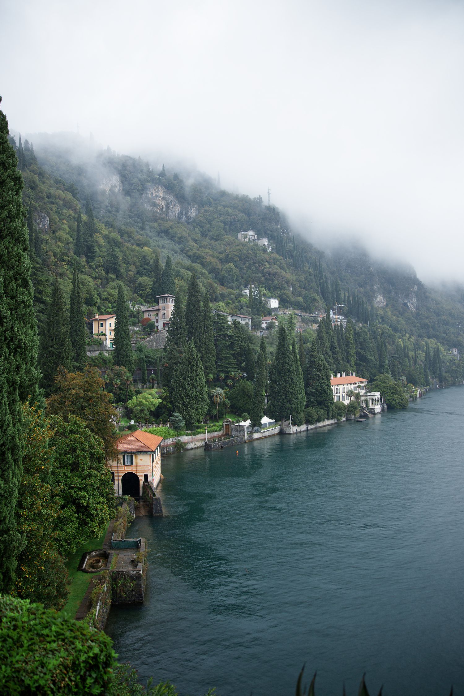 Misty lakeside villas and cypress trees line the shores of Lake Como in Italy on a foggy day.