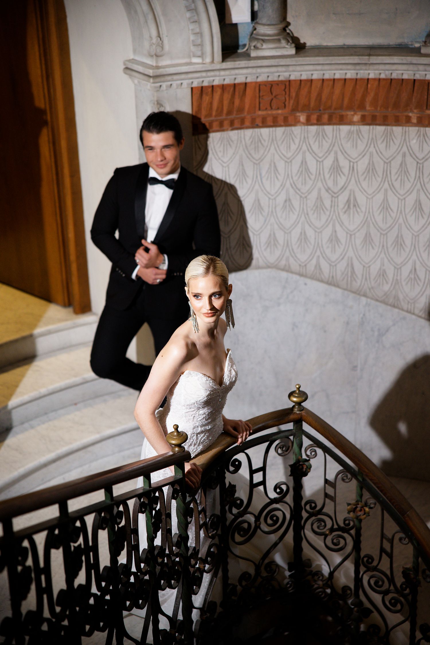 Wedding couple pose on an elegant curved staircase with ornate iron railings and white walls.