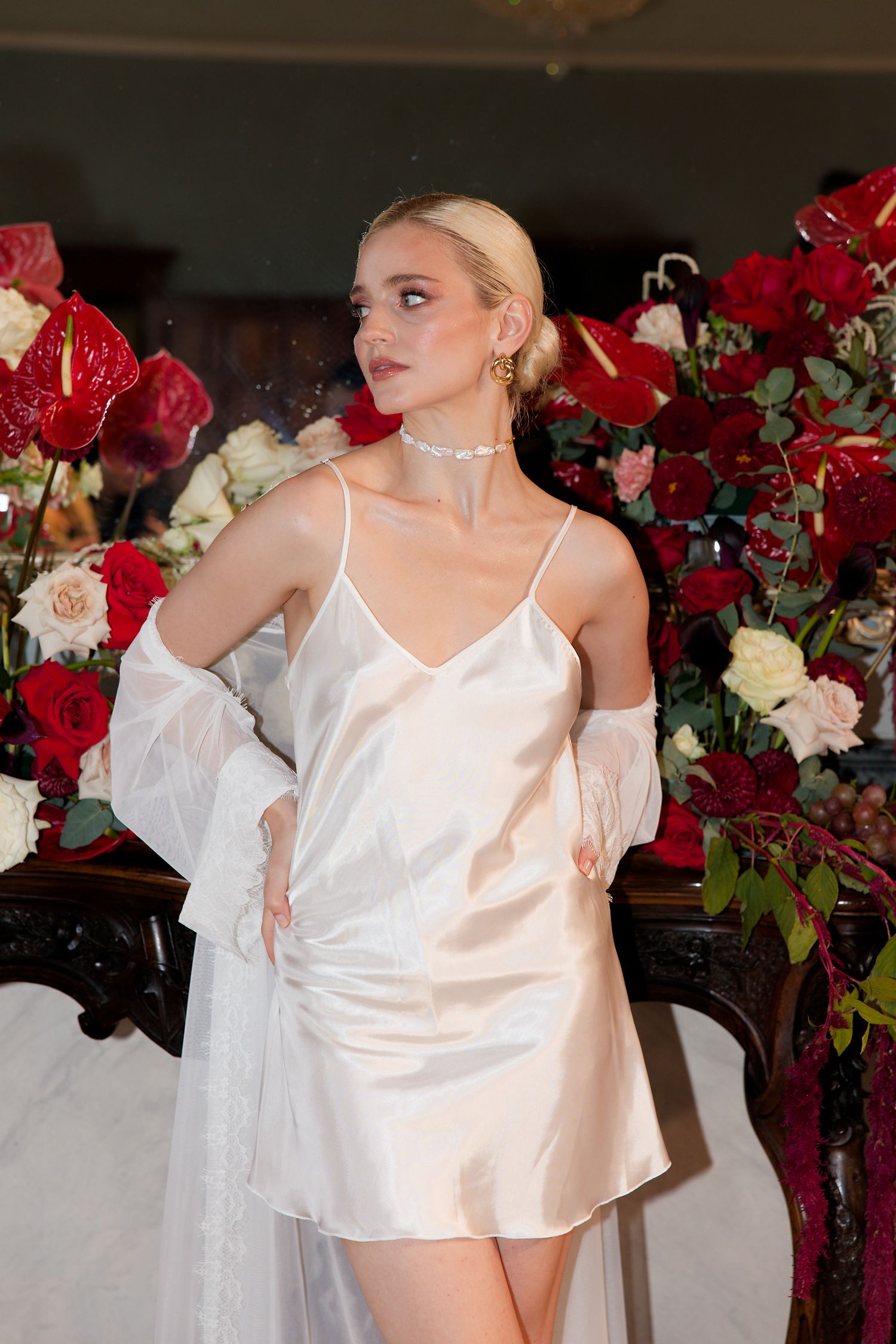 A woman in white silk dress and sheer sleeves poses elegantly against a backdrop of red and white roses.