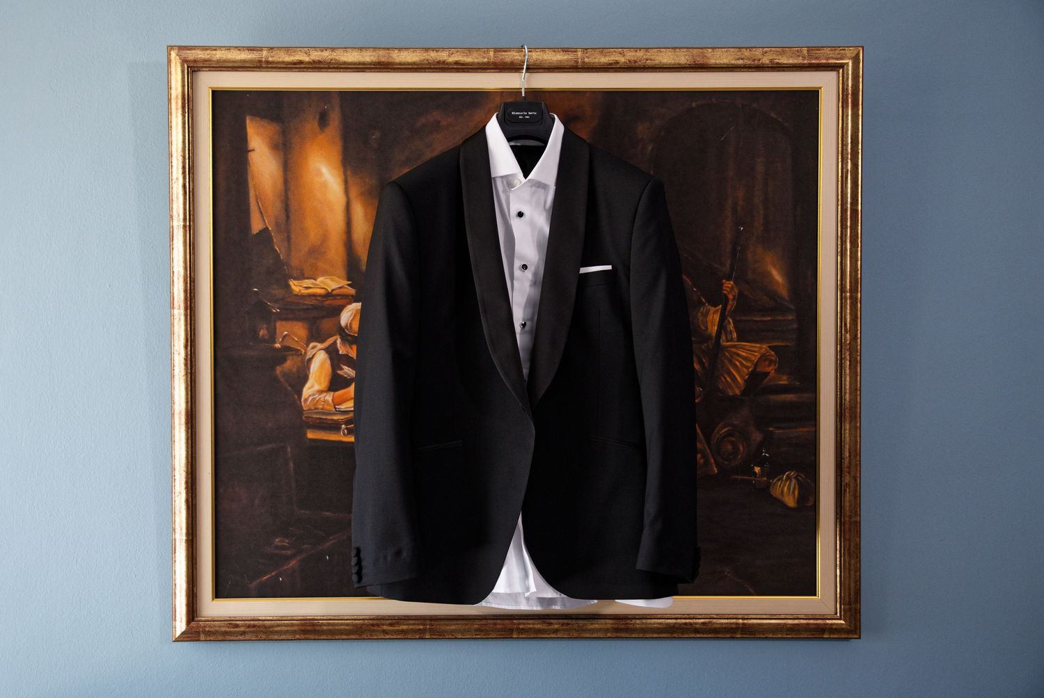 A black formal tuxedo jacket with white dress shirt displayed in an ornate gold frame against a blue wall.