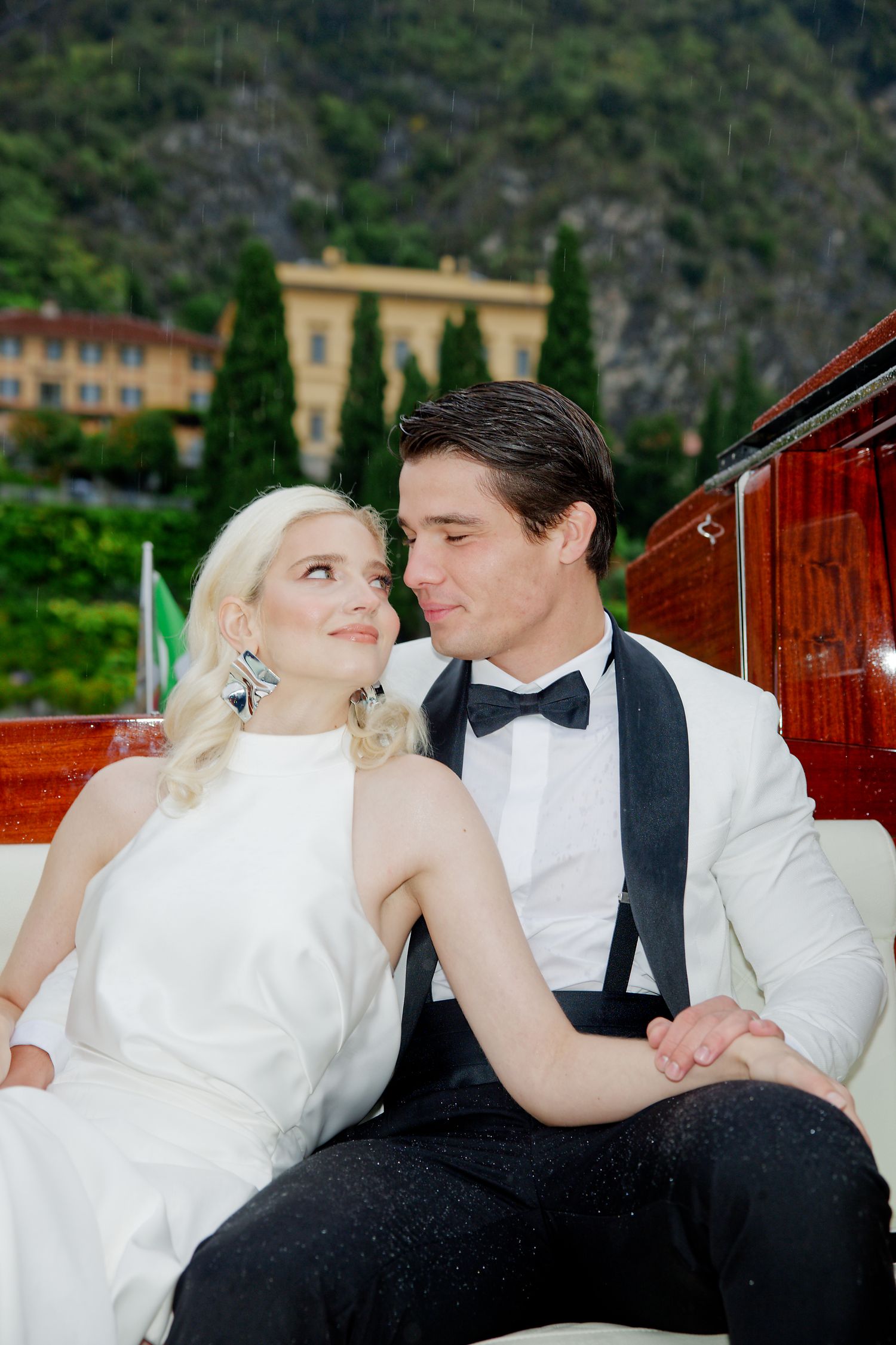 A couple in formal wedding attire shares an intimate moment on a luxury wooden boat with Italian villas in the background.