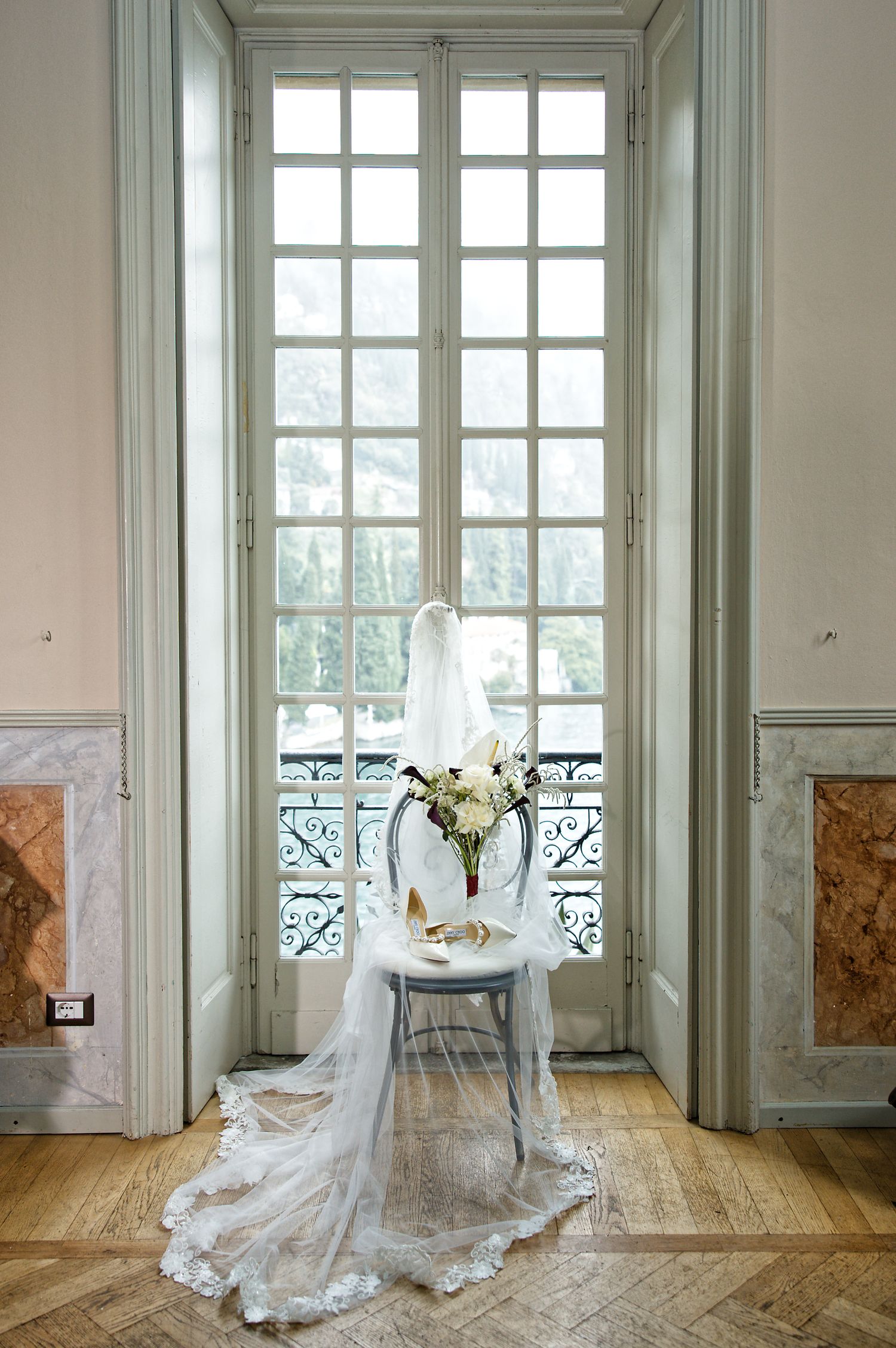 Wedding dress and bouquet elegantly displayed on white chair by French windows.