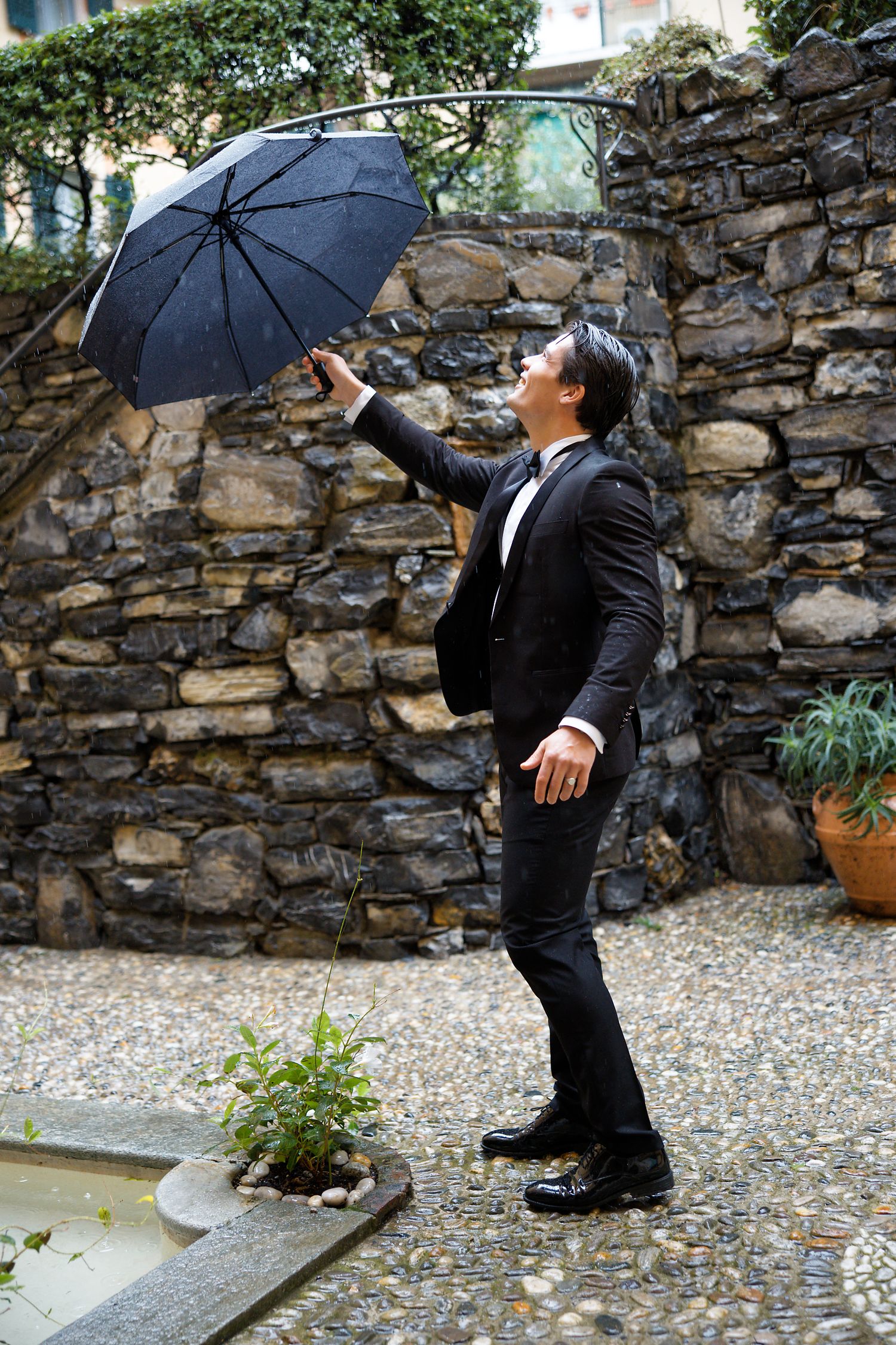 A person in a black suit poses playfully with an umbrella against a rustic stone wall in an outdoor garden setting.