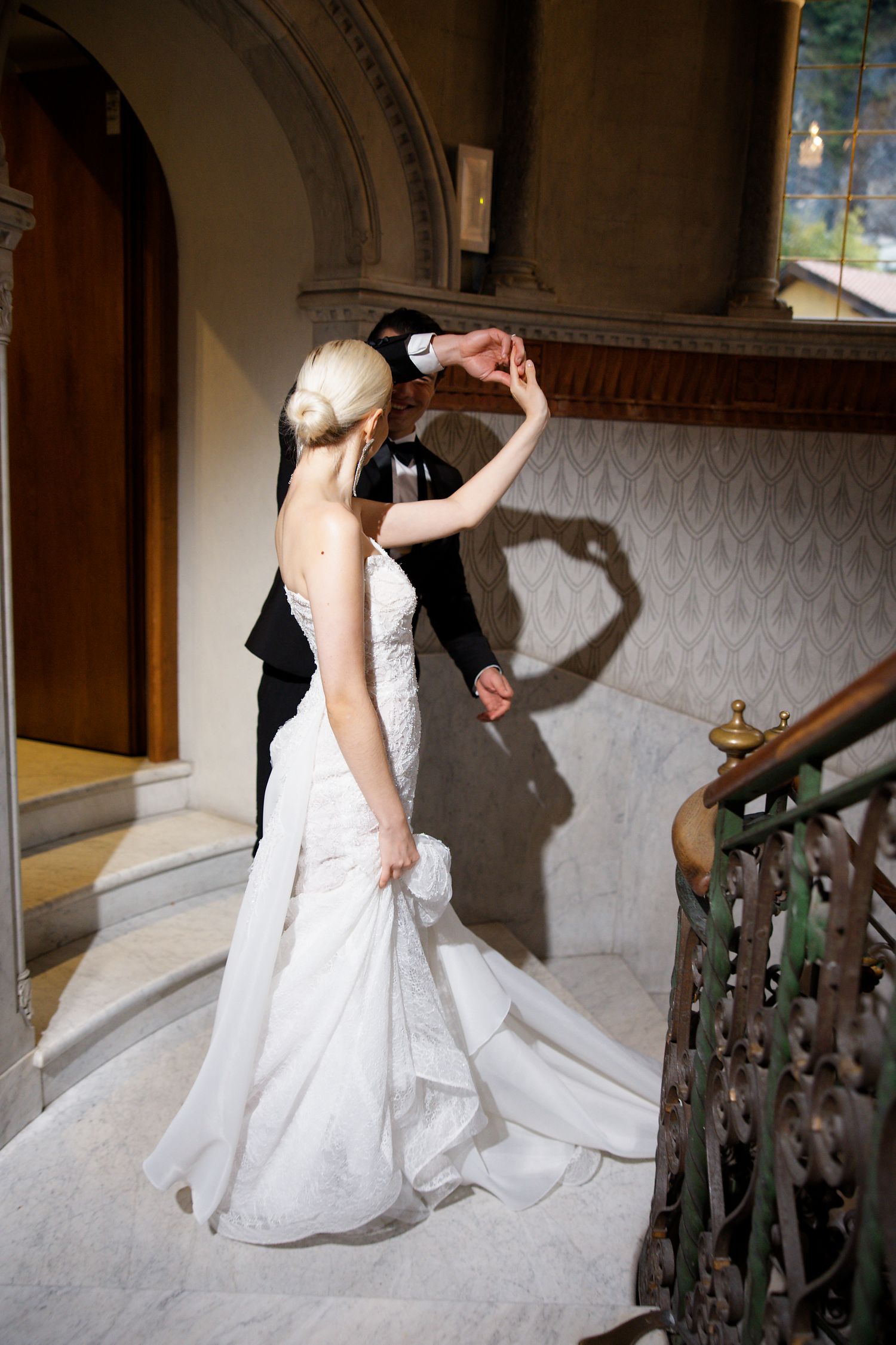 A wedding couple in elegant attire dance on a grand staircase with ornate architectural details.