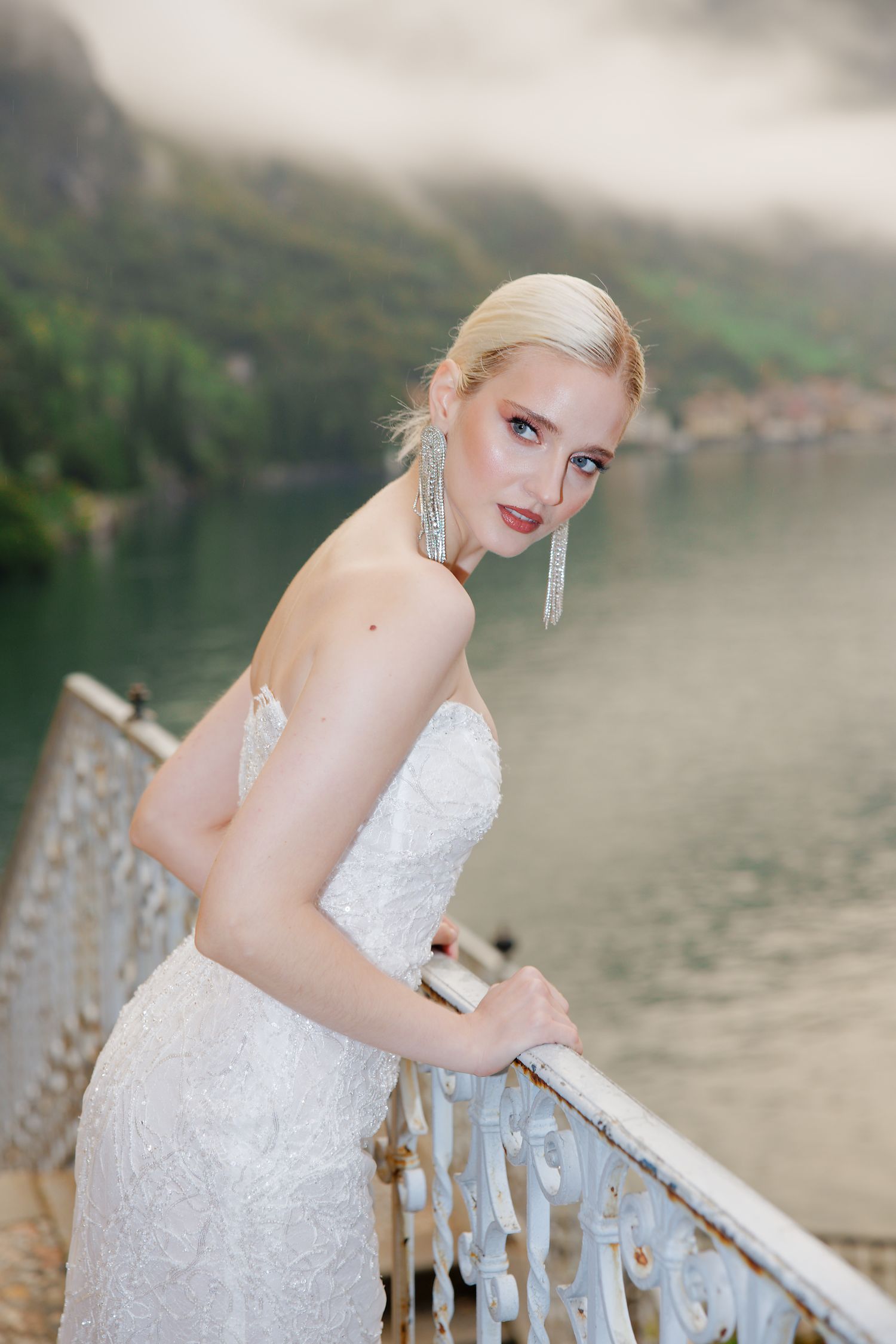 A bride in a white lace gown poses on an ornate balcony overlooking a misty lake.