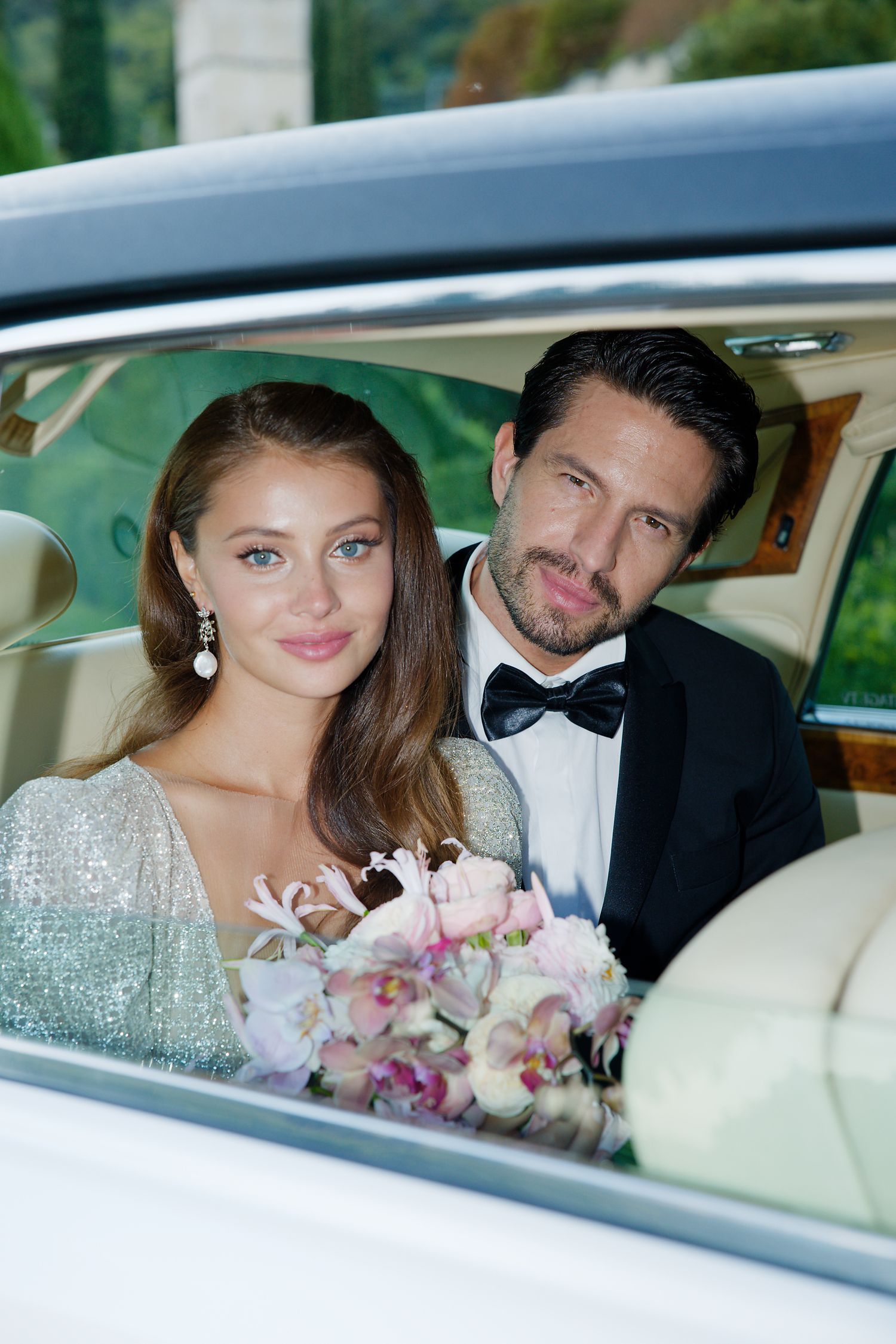 Newlyweds pose together in a classic car with a pink and white bridal bouquet.