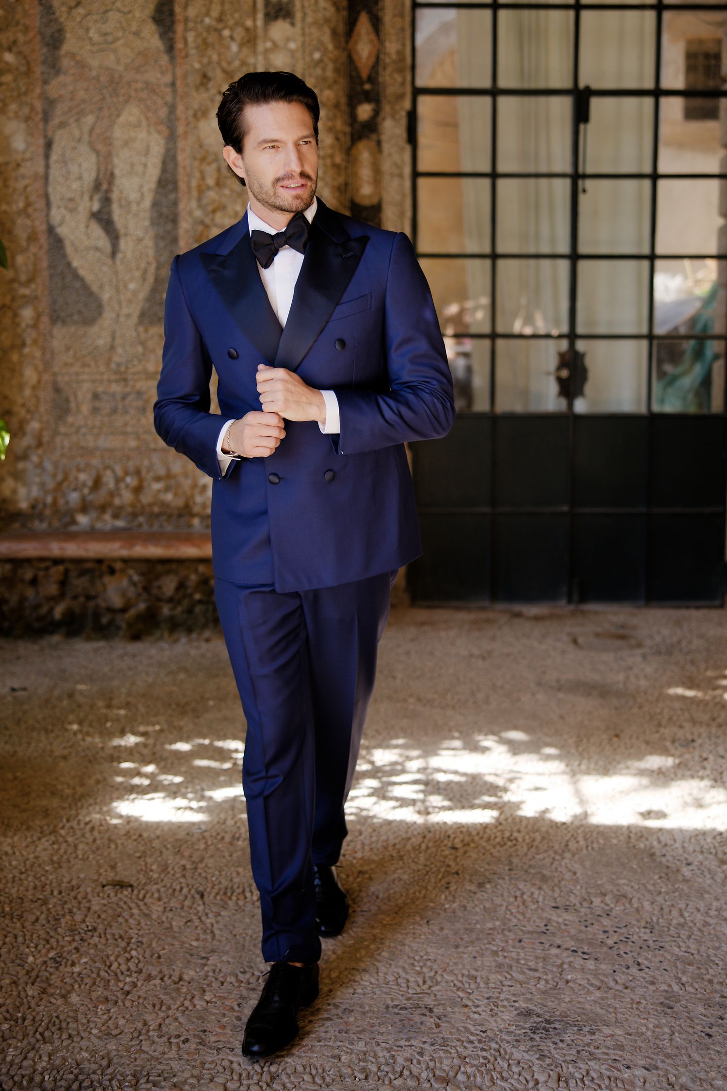A groom wearing a navy blue tuxedo with black lapels poses in an elegant setting.